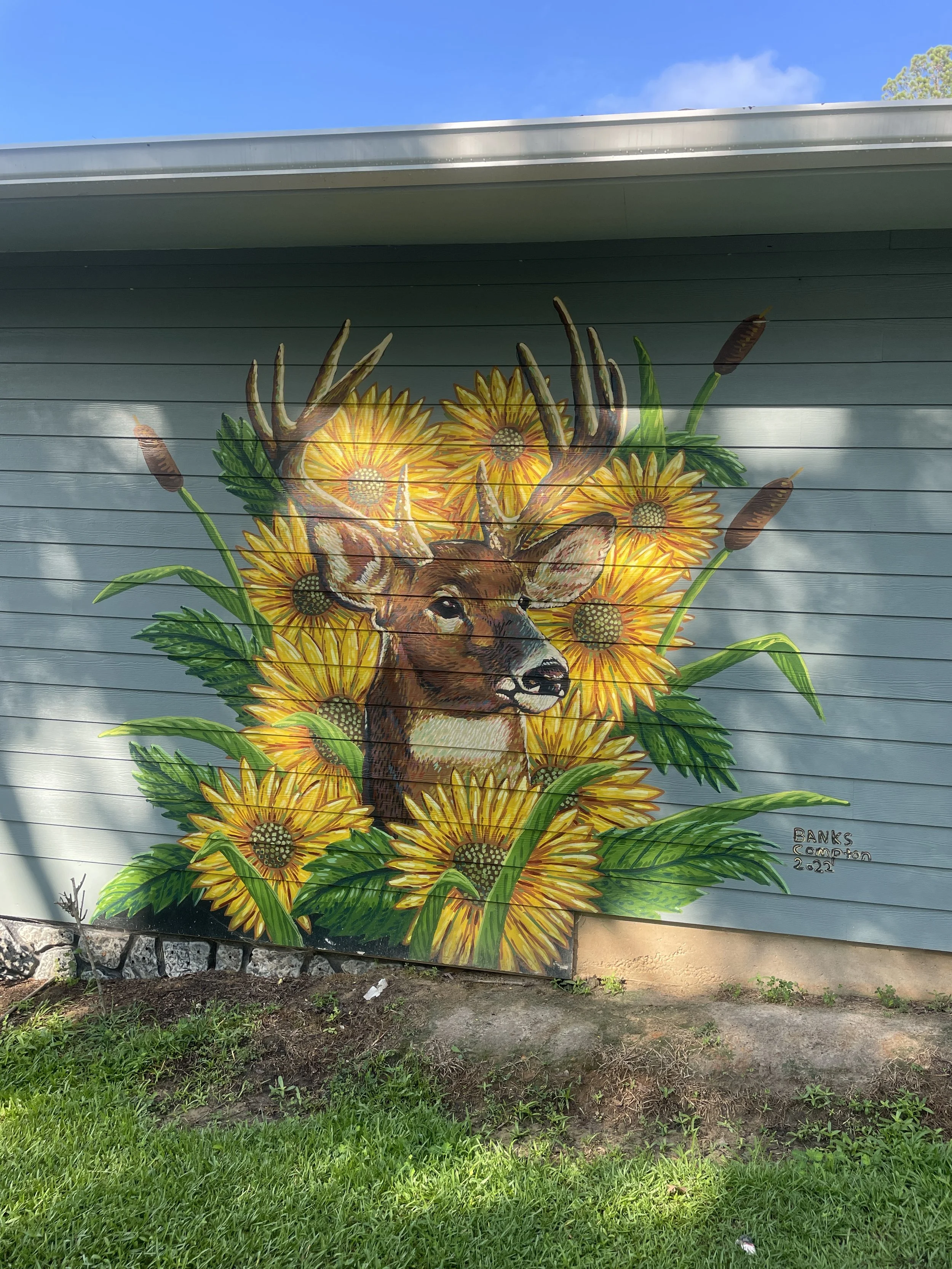 Residential mural by Banks Compton in Demopolis, Alabama, featuring a white-tailed deer surrounded by oversized yellow flowers, cattails, and foliage.