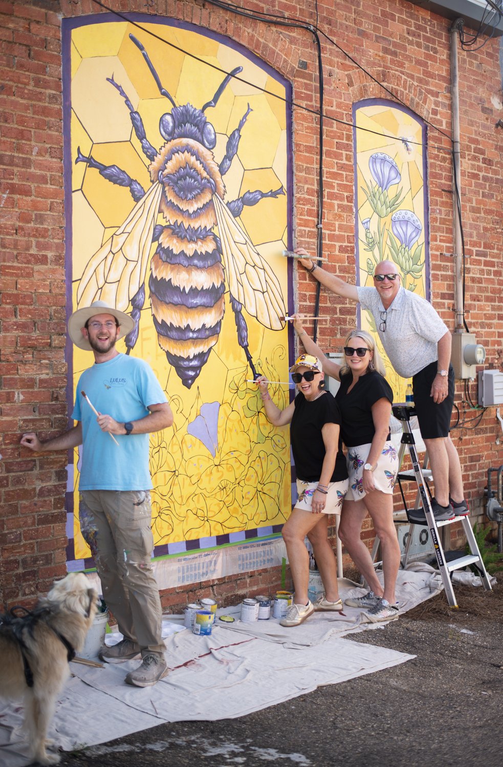 Group of four people painting a large mural on a brick wall featuring a bee and flowers, with a dog sitting nearby.