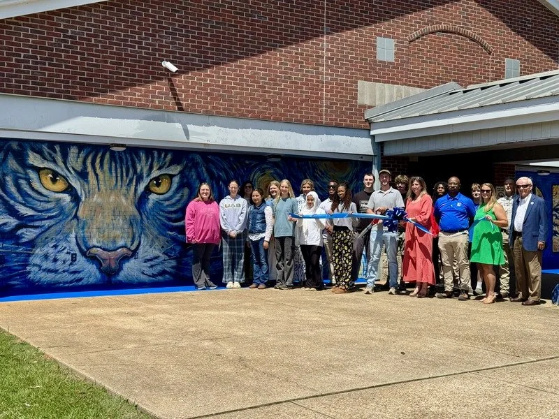 Group of people standing in front of a mural of a tiger's face, which they are about to cut with a large ceremonial scissors during an outdoor event.