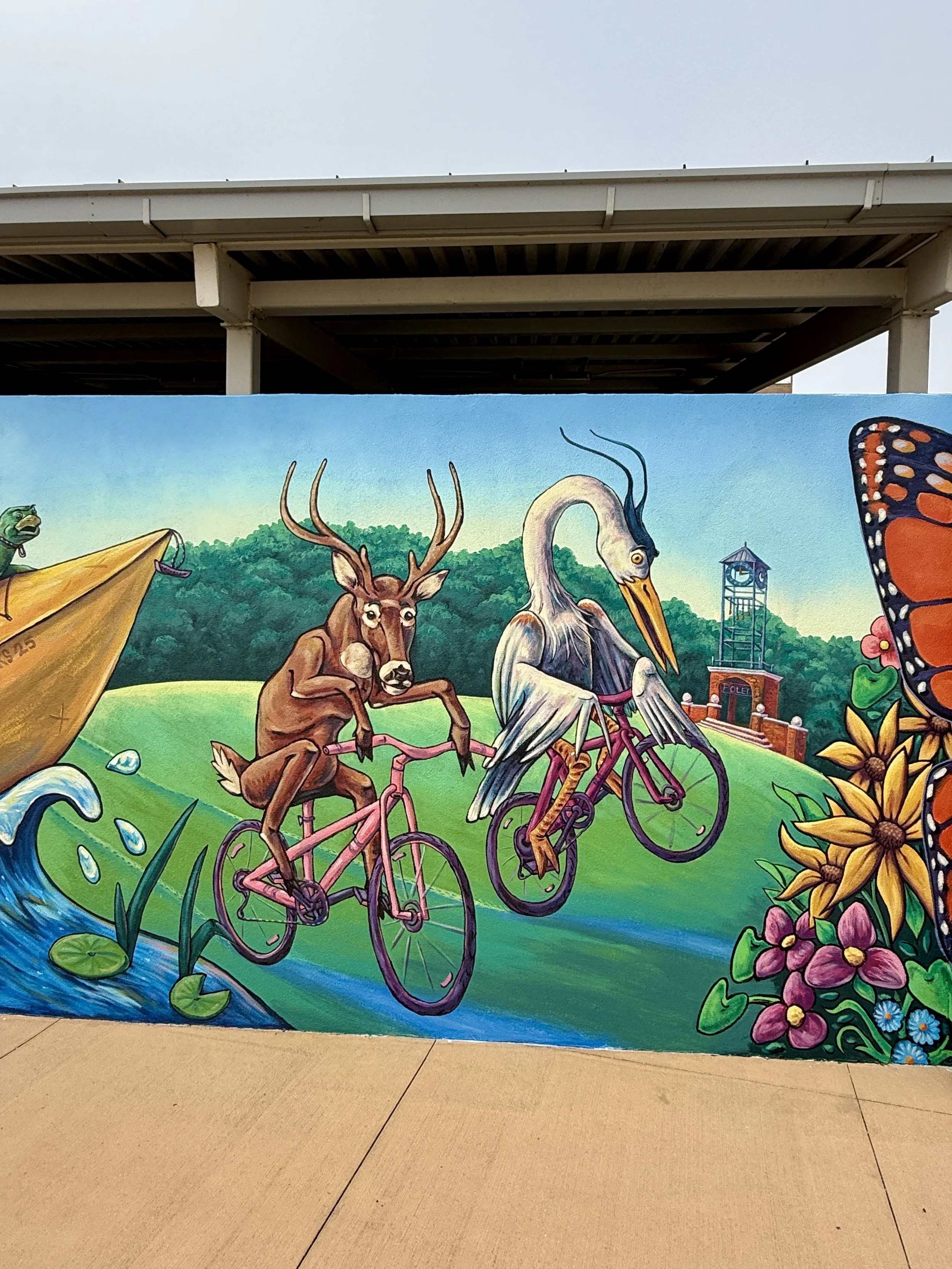 Colorful mural painting of various animals riding bicycles, with a river, greenery, and a tower in the background.