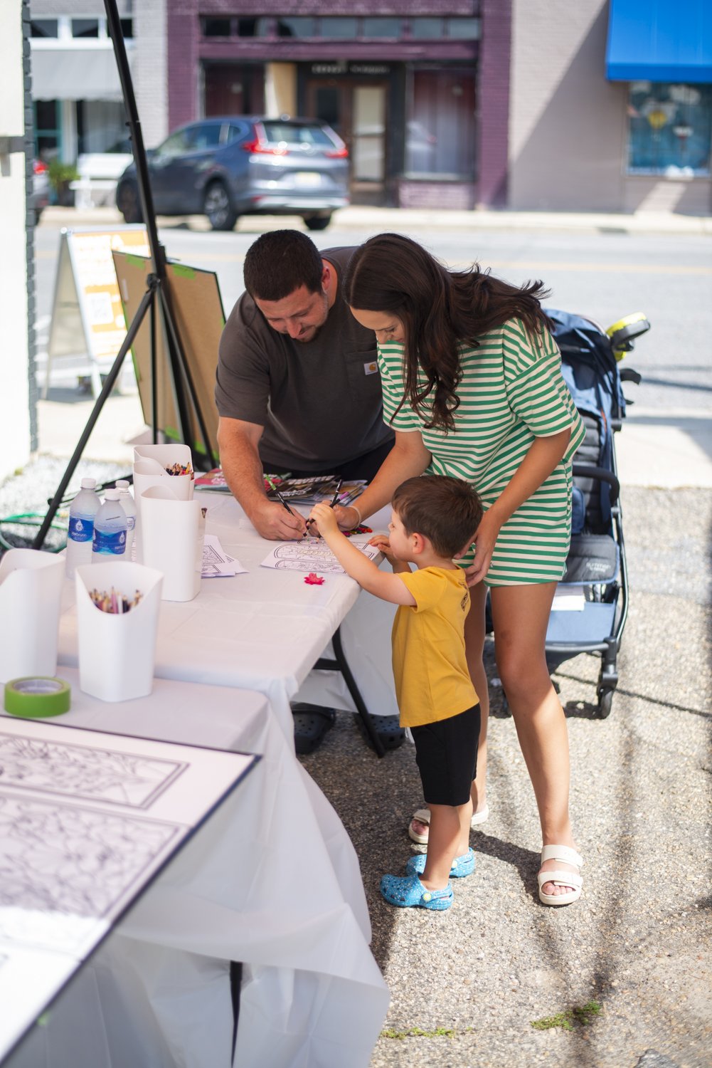 A family of three, including a young boy, a woman wearing a striped dress, and a man, gathered around a table outdoors on a city sidewalk. The man is helping the boy with coloring or drawing on a paper while the woman watches. The table is set with c