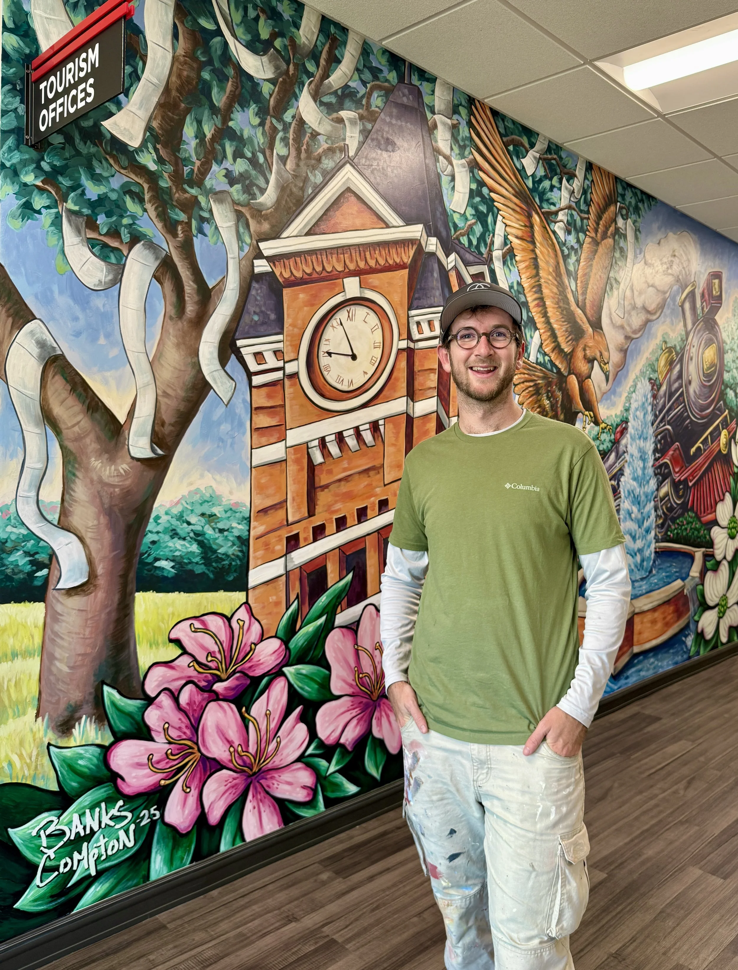 A young man wearing glasses, a gray cap, a green Columbia shirt, and white paint-splattered pants standing in front of a colorful mural inside a building. The mural depicts a tree with paper scrolls, a clock tower, pink flowers, an eagle, and a train.
