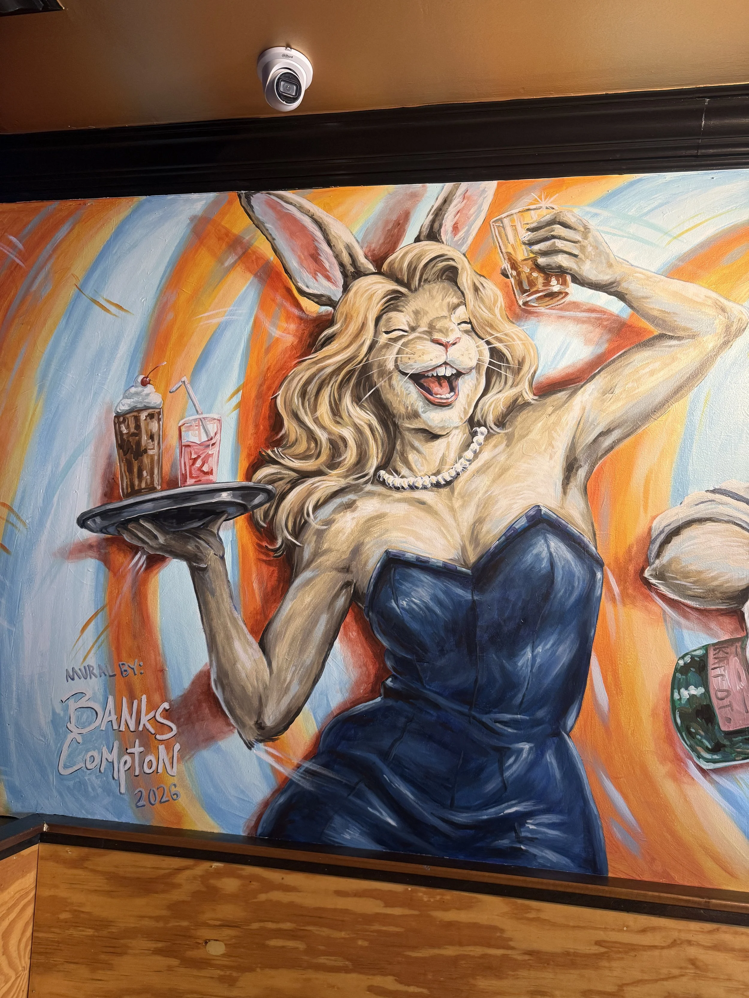 A colorful mural of a joyful anthropomorphic lion with long blond hair, wearing a blue dress, holding drinks on a tray, and wearing a pearl necklace, with vibrant orange, blue, and rainbow background.
