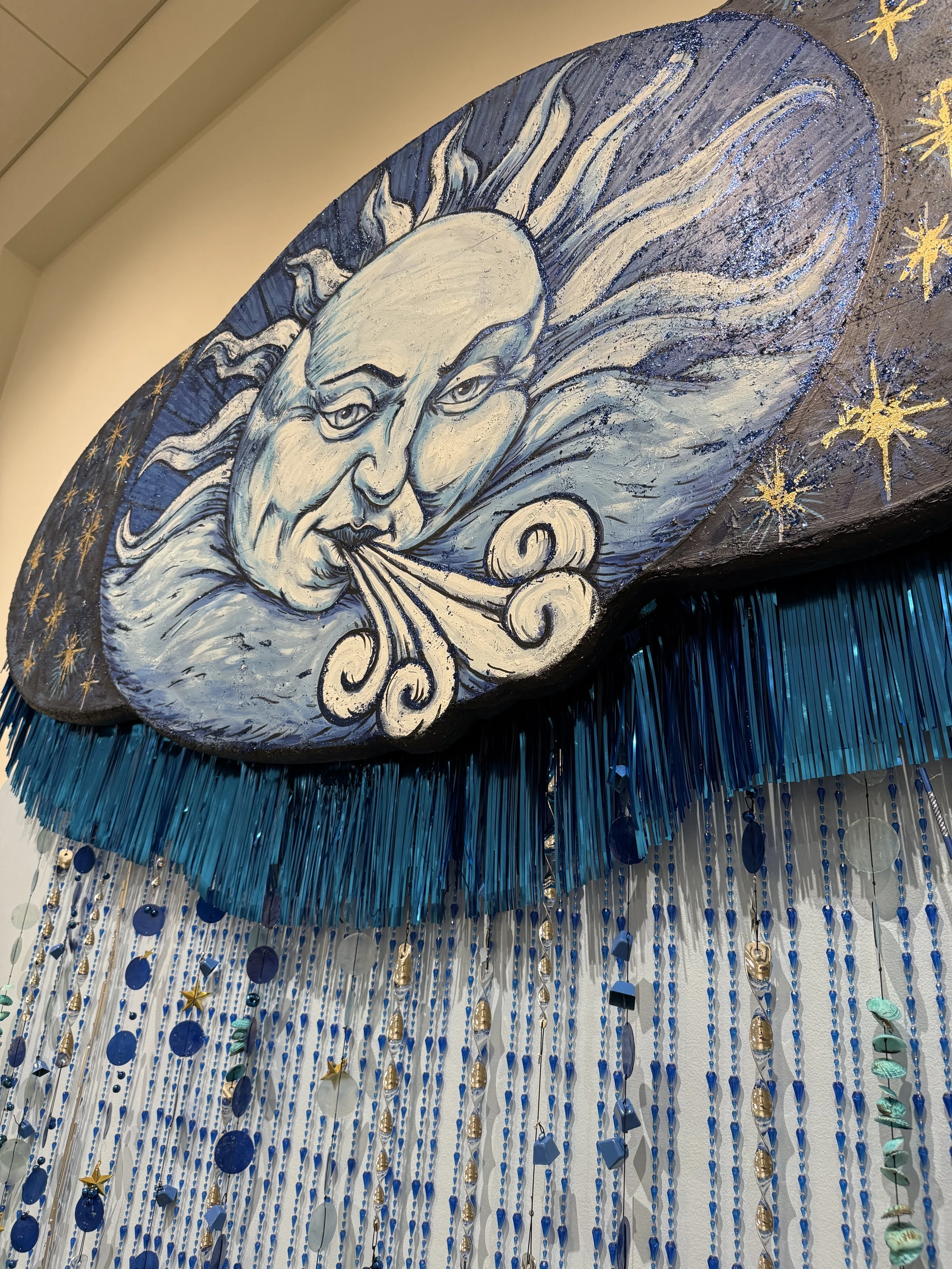 Close-up of Banks Compton's Color + Sound II sculpture showing the painted wind face, stars, blue fringe, beads, shells, and hanging sound elements.
