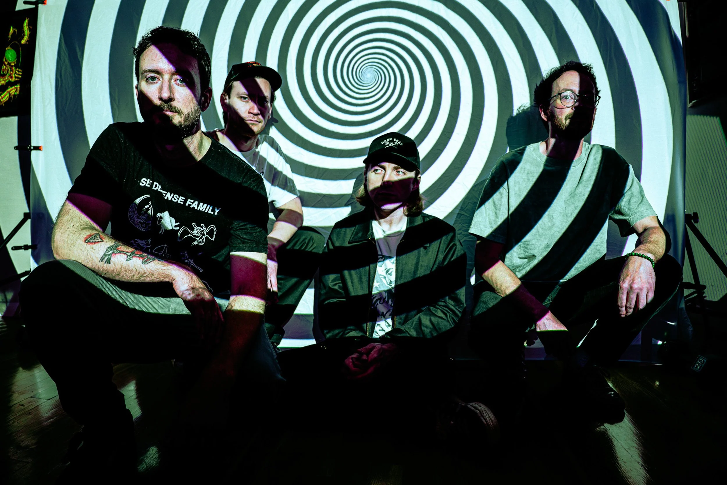 Four men sitting in front of a spiral hypnotic pattern projected on a wall.