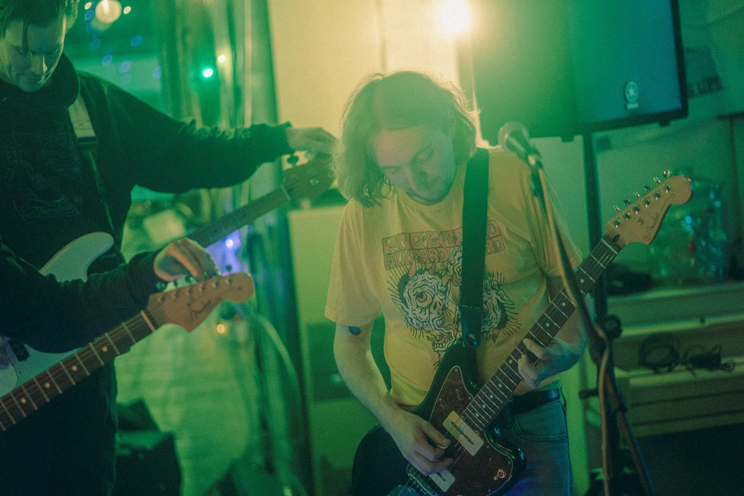 Two musicians playing electric guitars in a dimly lit room with green lighting. One musician is wearing a yellow T-shirt with a graphic design, and the other person is partially visible wearing a black hoodie. There is a microphone stand nearby and a