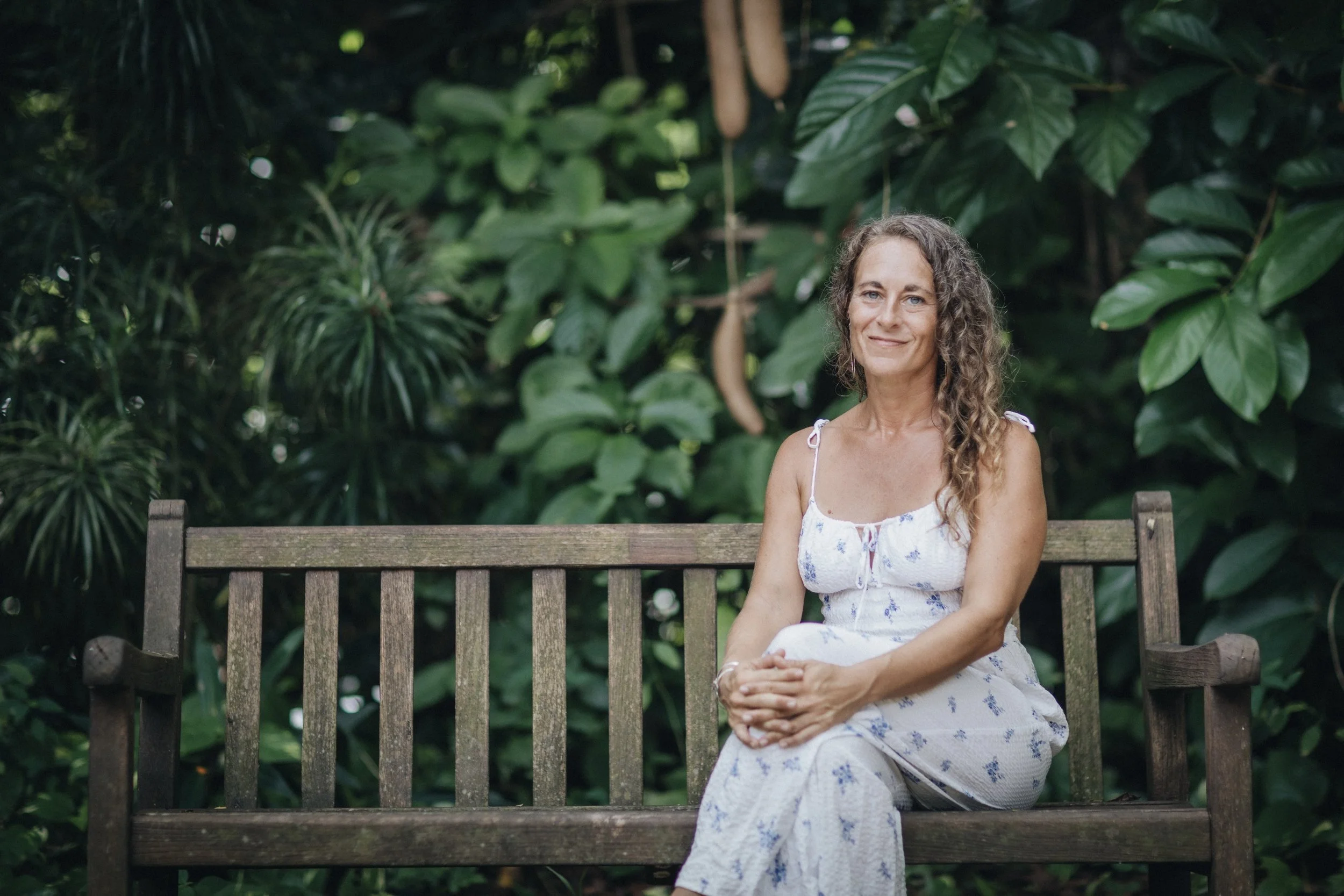 A woman with curly hair in a white dress with purple floral pattern sitting on a wooden bench in front of lush green foliage.