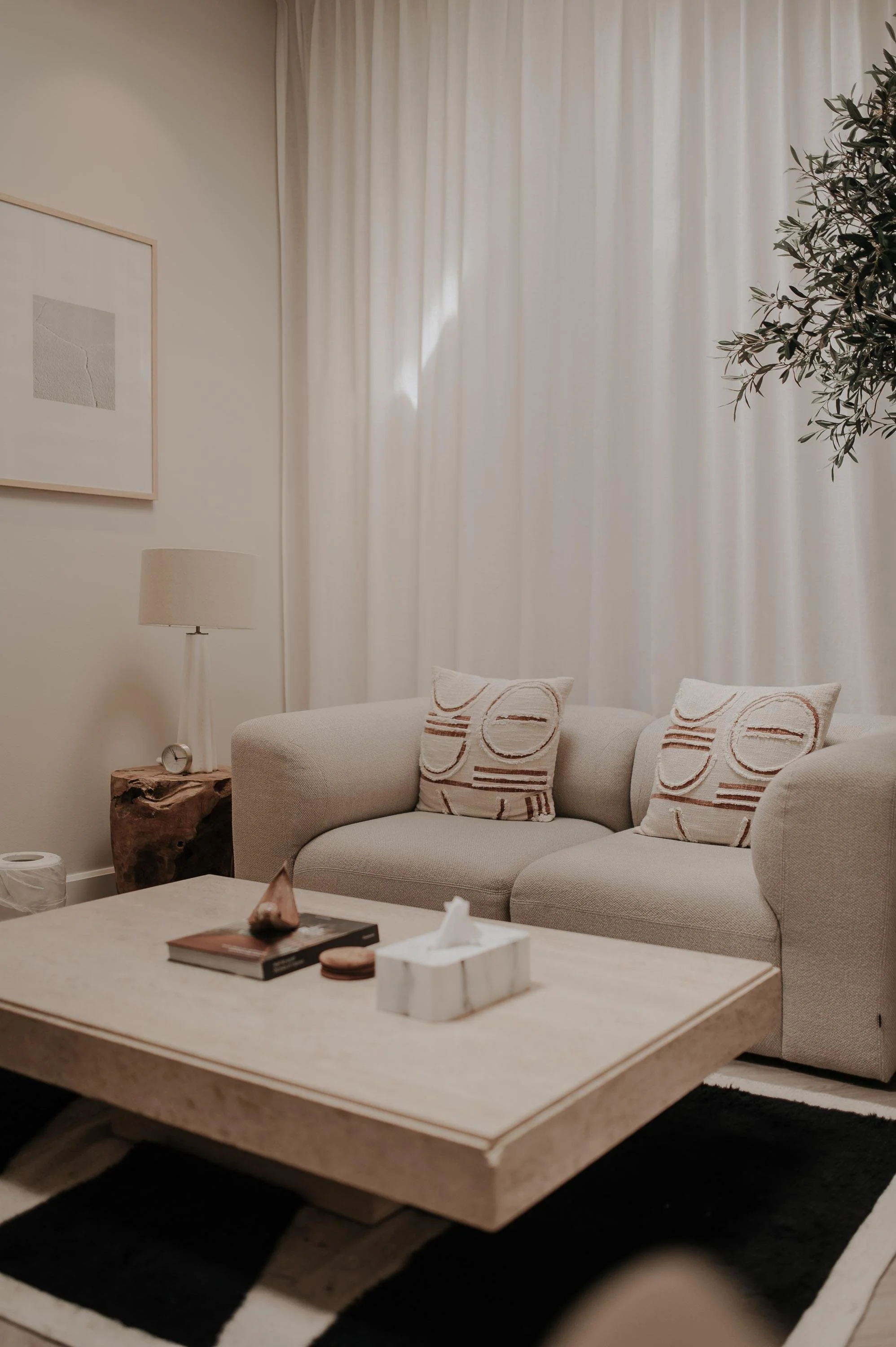 Woonkamer met beige bank, muurkunst, tafellamp op houten tafel, decoratieve kussens, witte gordijnen, planten en zwarte en witte vloerkleed.