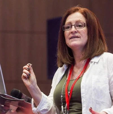 Woman with glasses and shoulder-length hair speaking at a podium, holding a small object in her right hand, wearing a white jacket and red lanyard.
