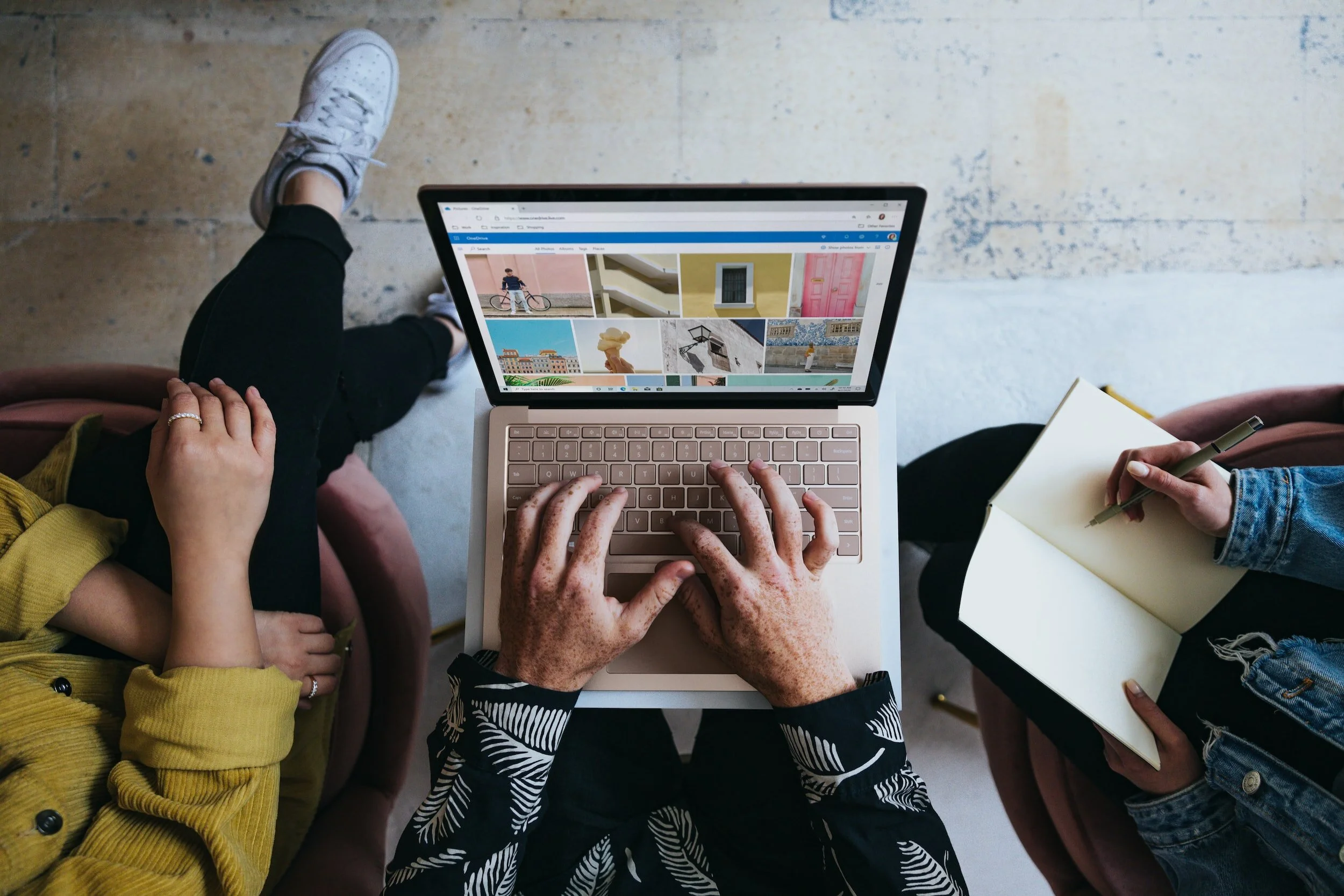 People sitting around a table with a laptop, one person typing, another with a notebook and pen, viewing and taking notes on a website showing various images of buildings and places.