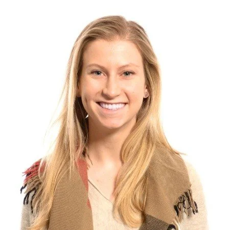 A young woman with long blonde hair smiling, wearing a tan and beige scarf with fringe details on a plain white background.