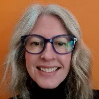 Close-up of a smiling woman with gray hair and glasses, against an orange background.