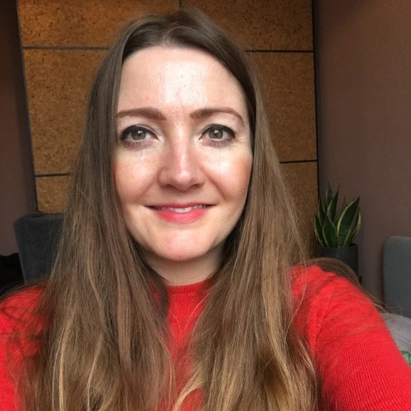 A smiling woman with long brown hair wearing a red sweater, sitting indoors near a potted plant and a wall with wooden panels.