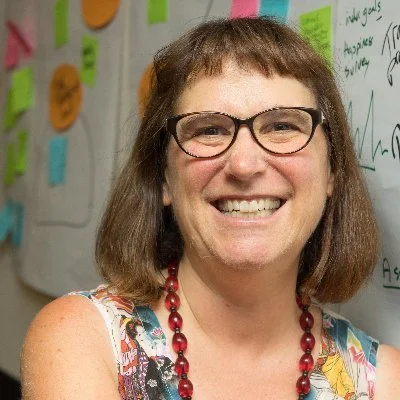 A smiling woman with glasses and shoulder-length brown hair, wearing a colorful sleeveless top and a red beaded necklace, standing in front of a whiteboard with colorful sticky notes and handwritten notes.