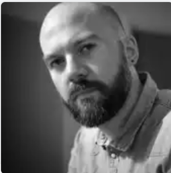 Black-and-white portrait of a man with a beard and short hair, wearing a button-up shirt, looking at the camera.