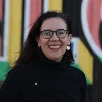 A woman with dark wavy hair, glasses, and pearl earrings smiling, standing outdoors in front of colorful graffiti.