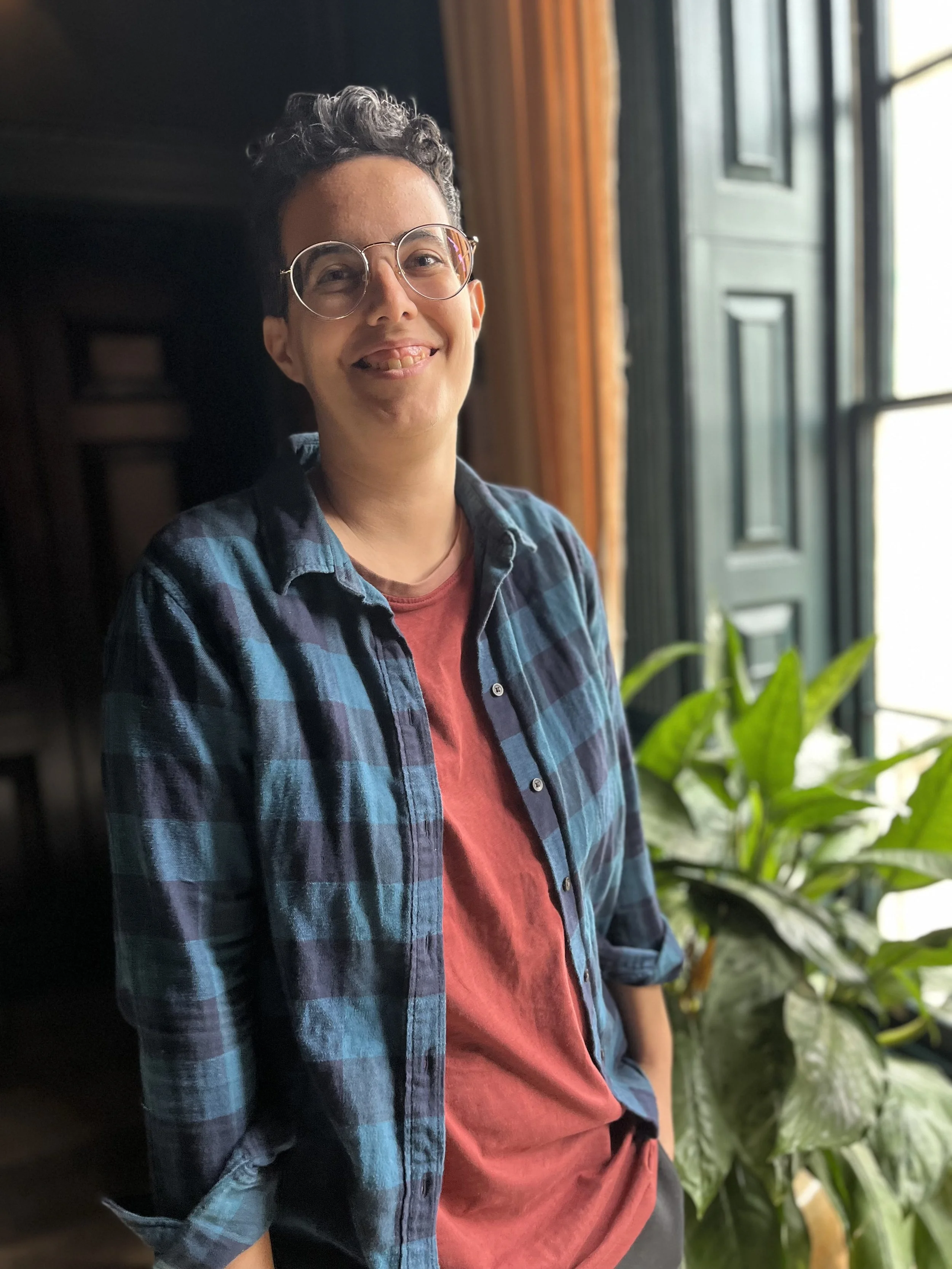 A person with short dark hair and glasses, smiling slightly with their tongue out, standing inside near a window with green plants in the background.