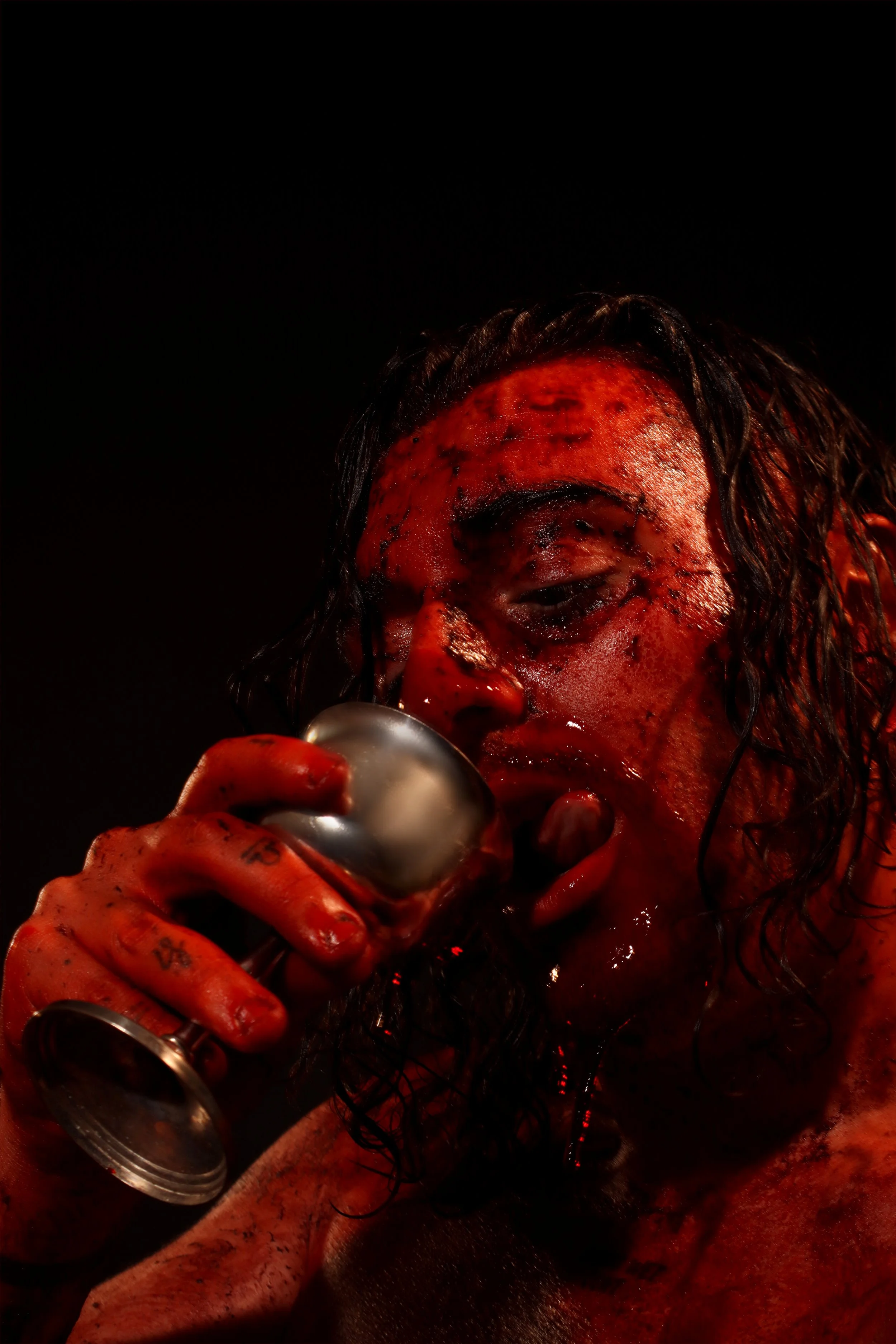 A person with long, wet hair and makeup resembling blood on their face, holding a goblet near their mouth.