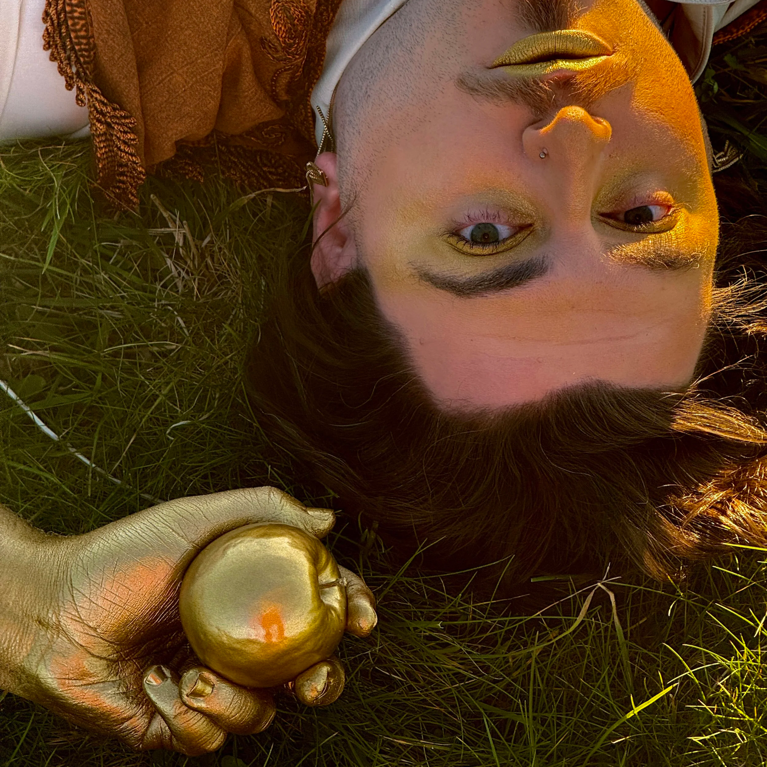 A person with brightly colored makeup lying on grass, holding a shiny gold apple. The person has gold eyeshadow, gold lipstick, and a nose piercing, with a golden hand holding the apple.