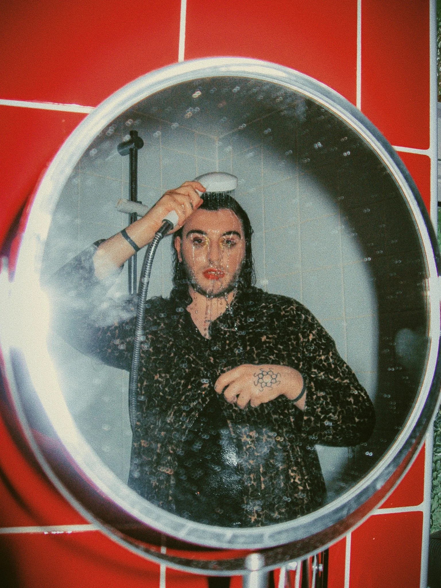 A person with dark hair, makeup, and tattoos taking a shower and rinsing their hair, seen through a bathroom mirror.