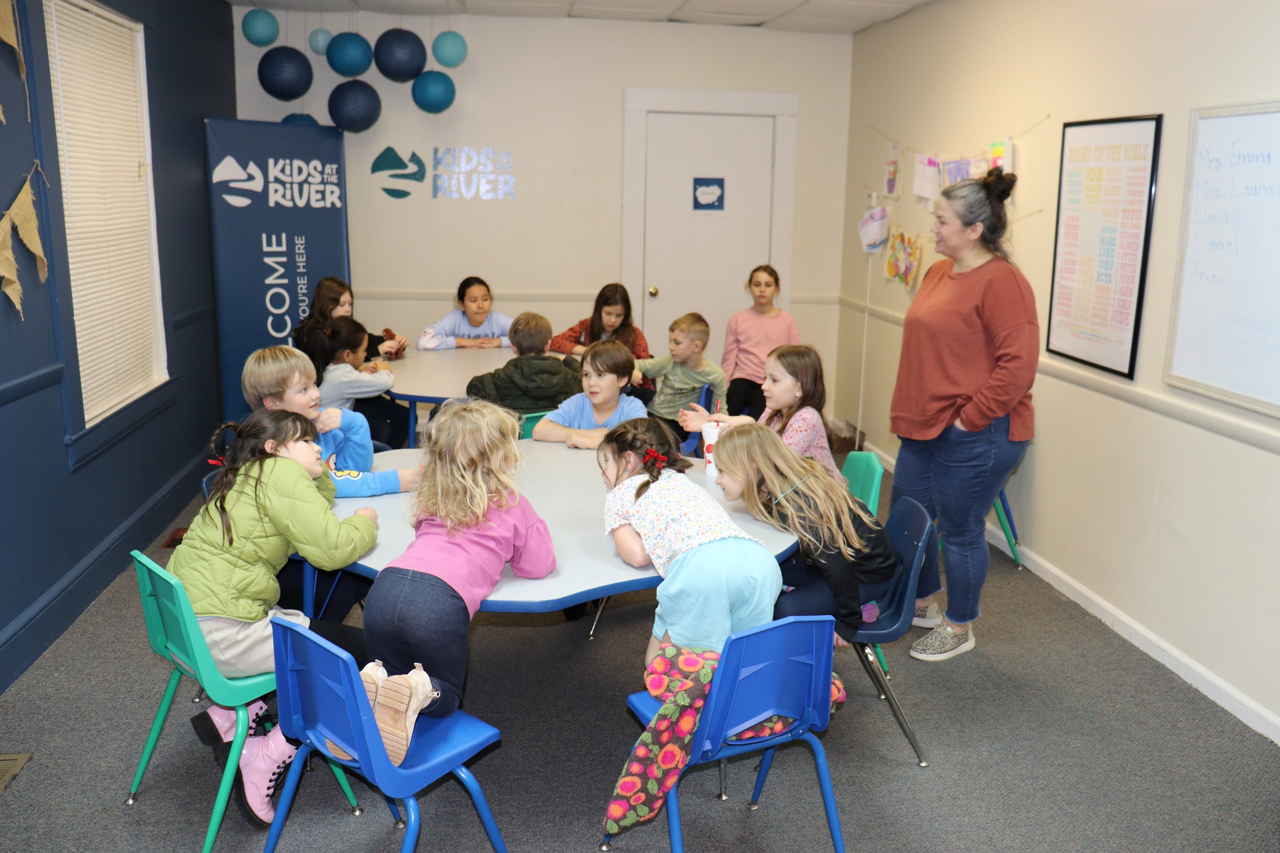 A woman is standing and speaking to a group of children sitting at a cafeteria table in a classroom. The children are listening attentively. There are decorations on the wall and banners indicating a Kids at the River program.