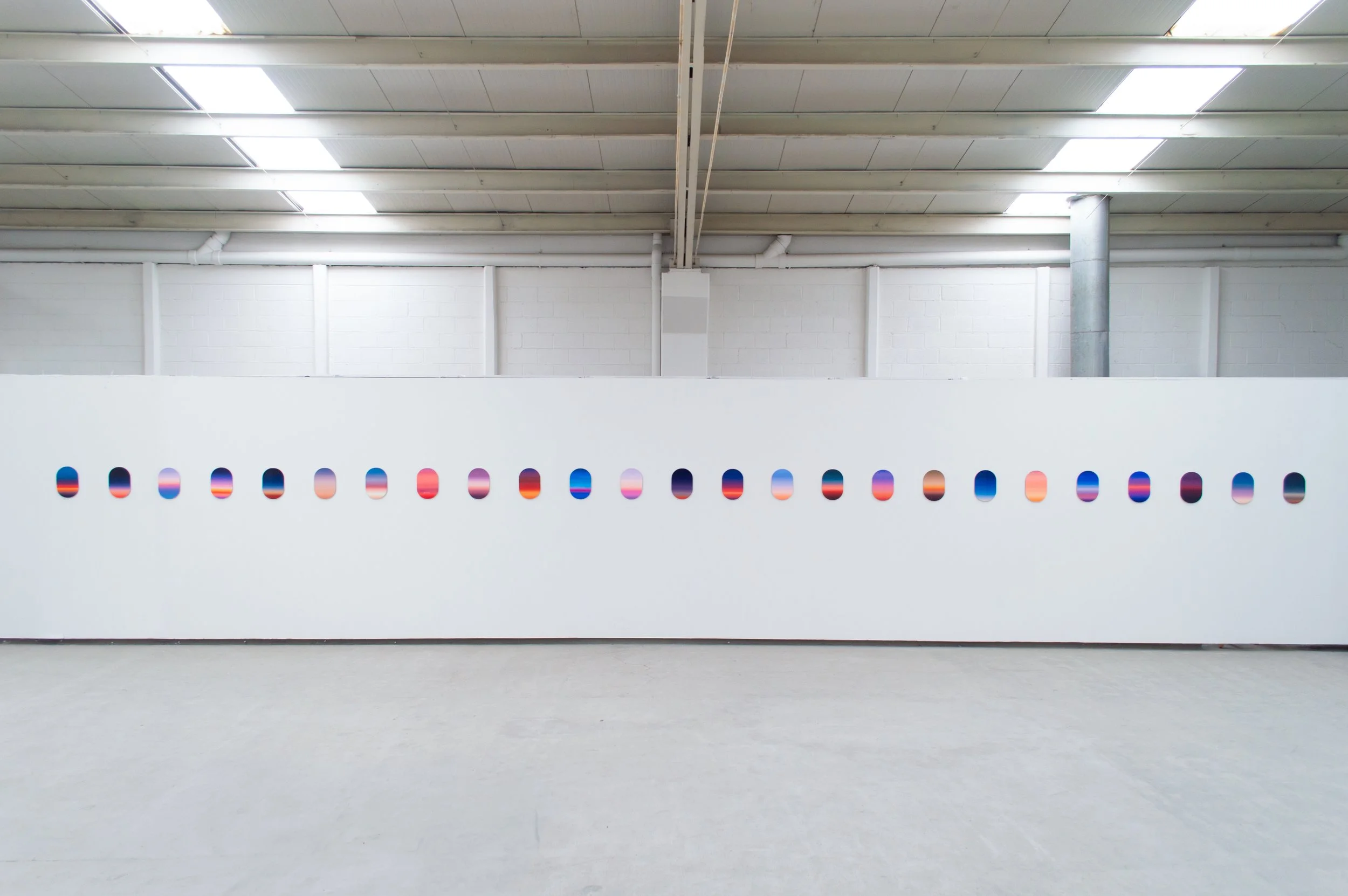 Artwork of a sunset or sunrise with multiple oval shapes mounted on a white gallery wall, arranged in a horizontal line, each displaying different gradient colors.