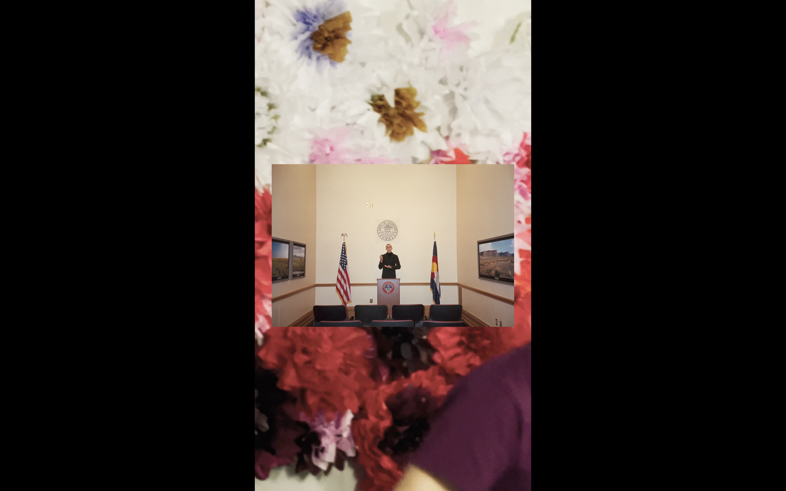 A man standing at a podium in a conference room with American and Colorado flags, flanked by seated chairs and framed pictures on the walls, viewed through a floral foreground. art video by swiss artist SEVERIN HALLAUER - consisting  of analog photos