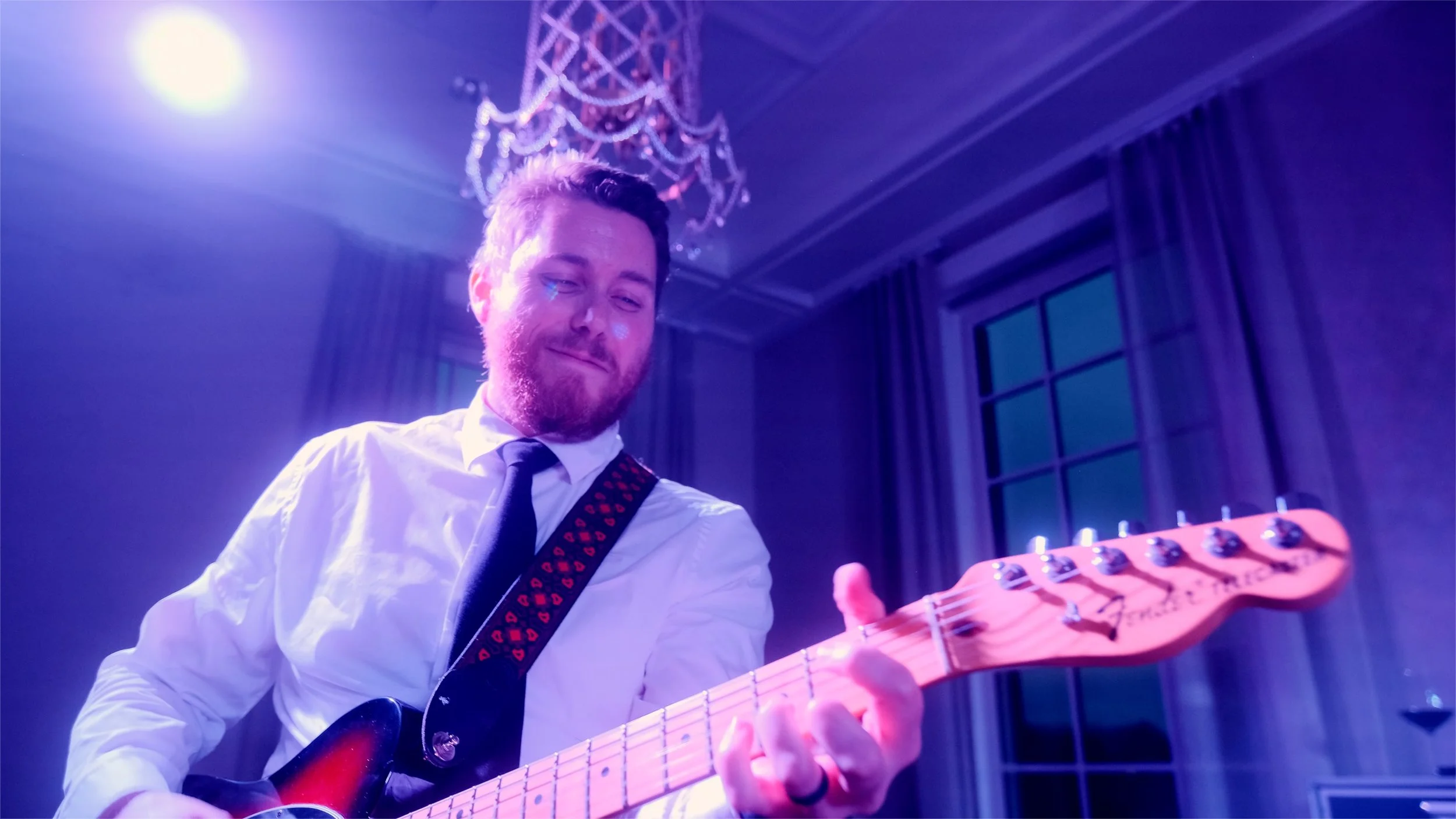 A man with a beard and short hair playing an electric guitar indoors, illuminated by purple stage lights, with a chandelier and large windows in the background.