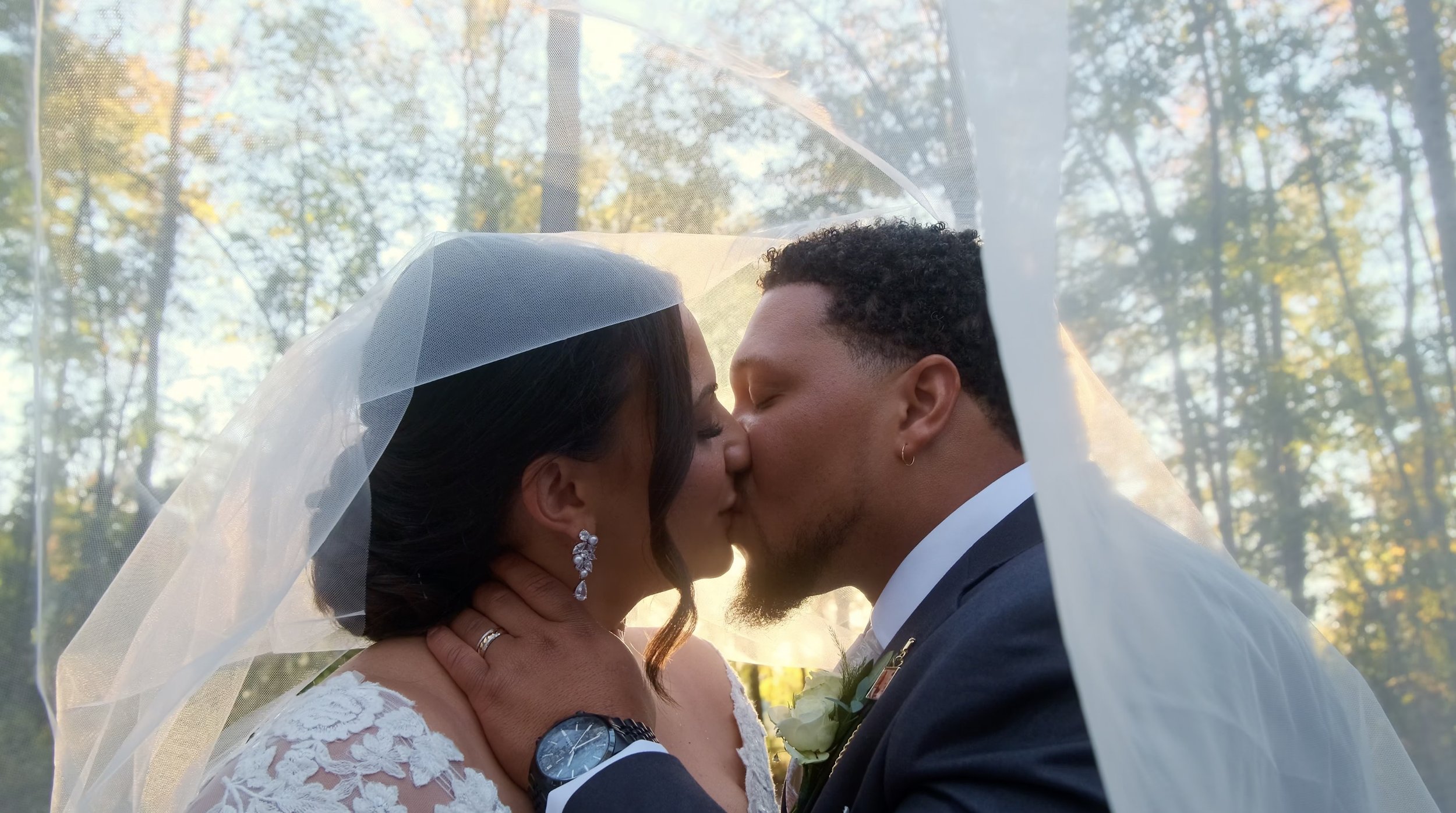 A bride and groom kiss under a wedding veil outdoors in a wooded area with autumn trees.