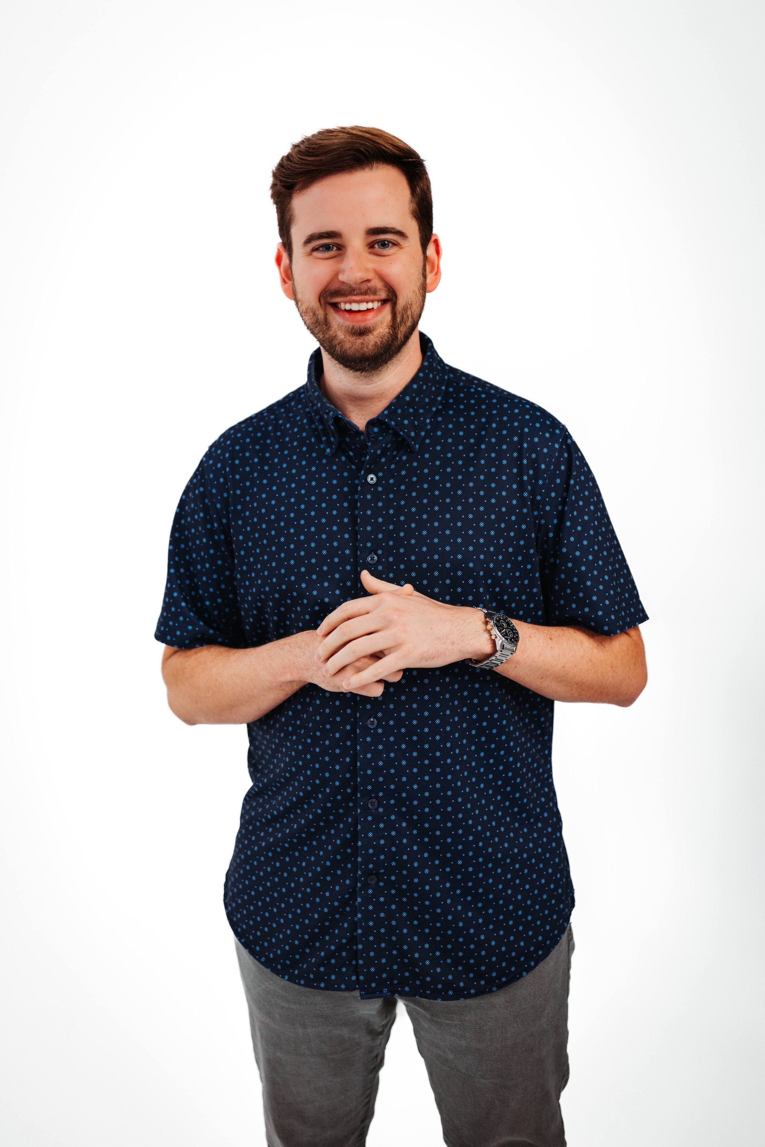 A young man with brown hair, beard, and blue eyes, smiling in front of a white background. He is wearing a navy blue shirt with small white polka dots, gray pants, and a wristwatch.