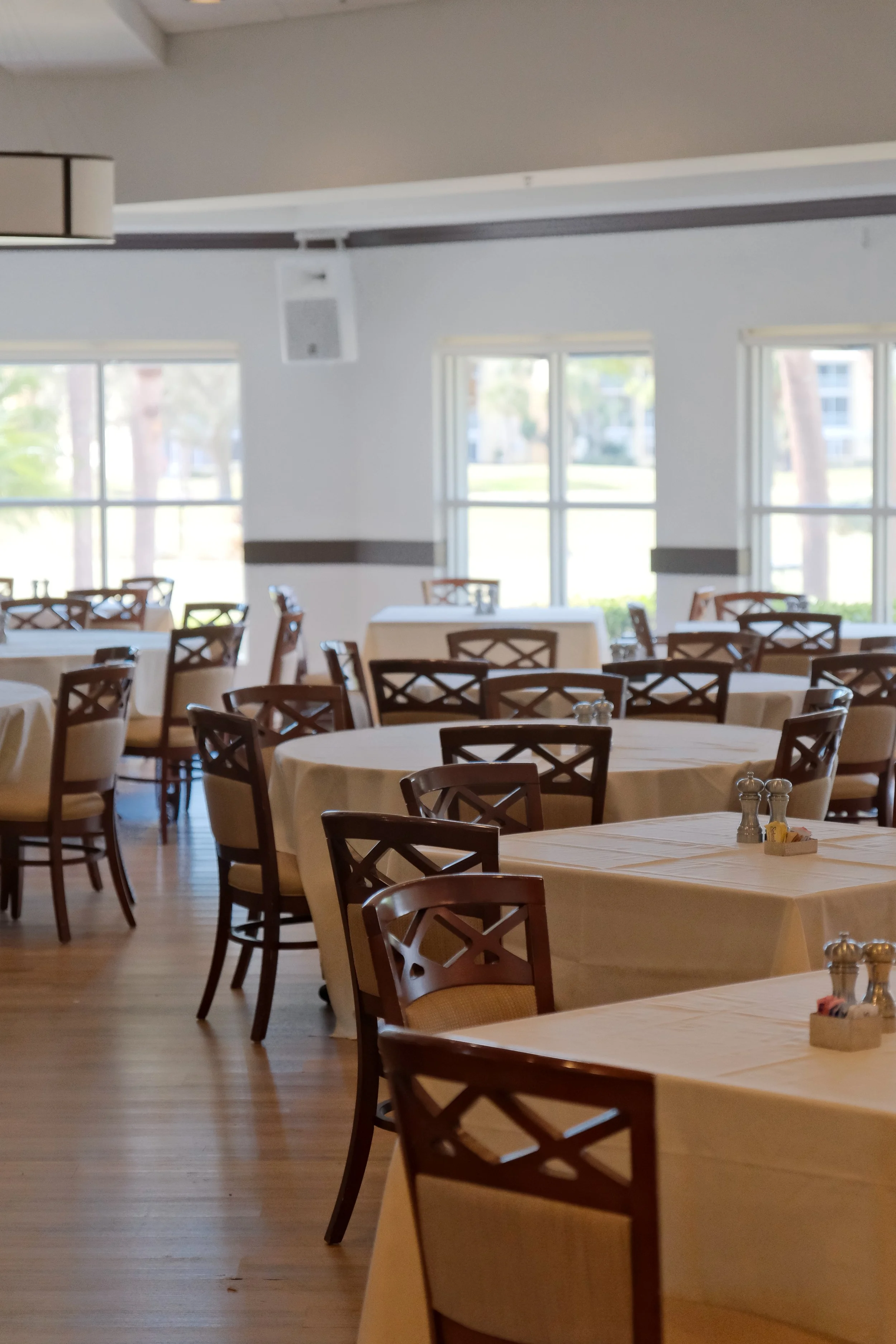 Empty restaurant dining area with round tables covered in white tablecloths and wooden chairs. Large windows let in natural light and show trees outside.