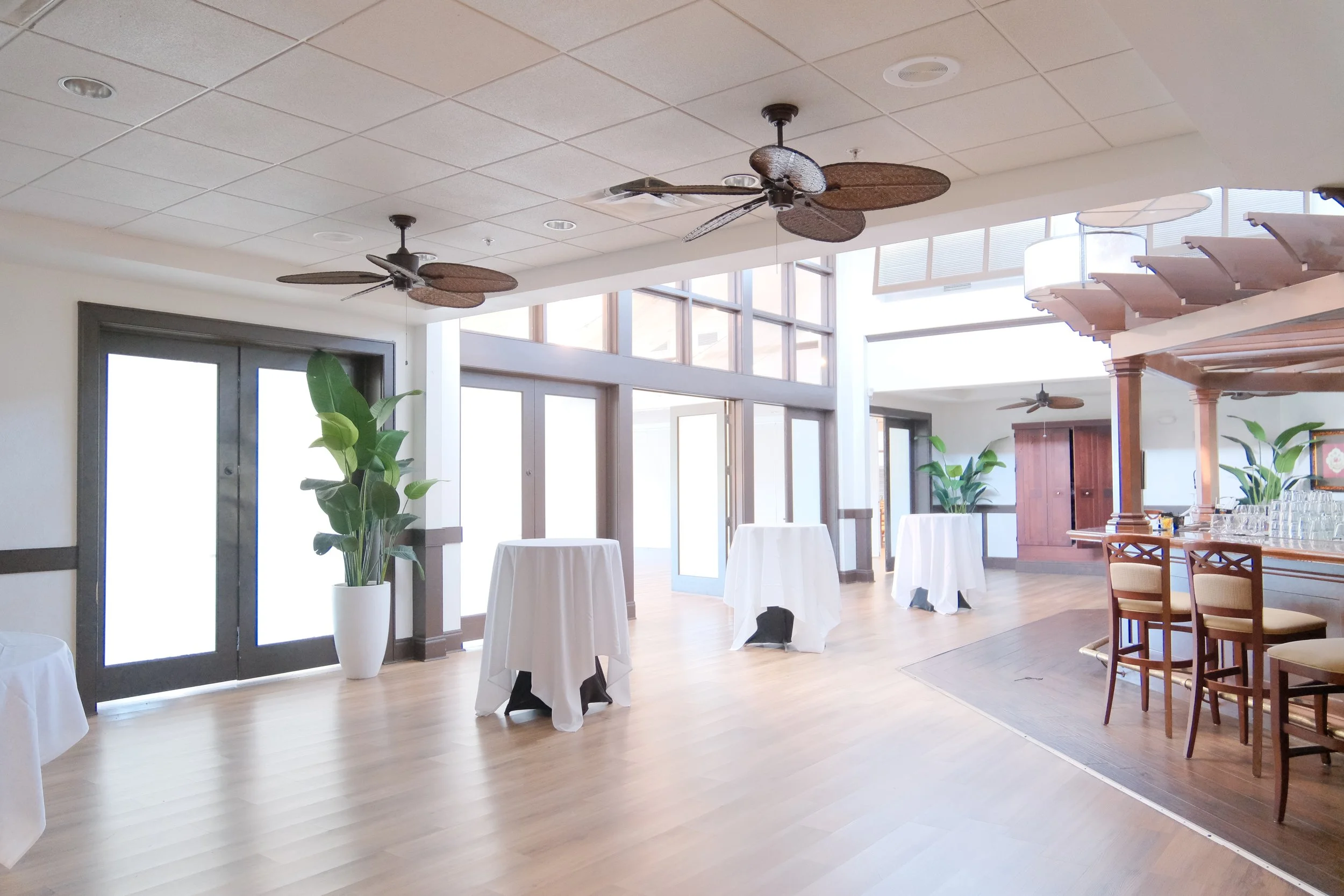 Bright, spacious indoor area with large glass windows, wooden floors, ceiling fans, green potted plants, and tables covered with white tablecloths, likely set up for an event or reception.