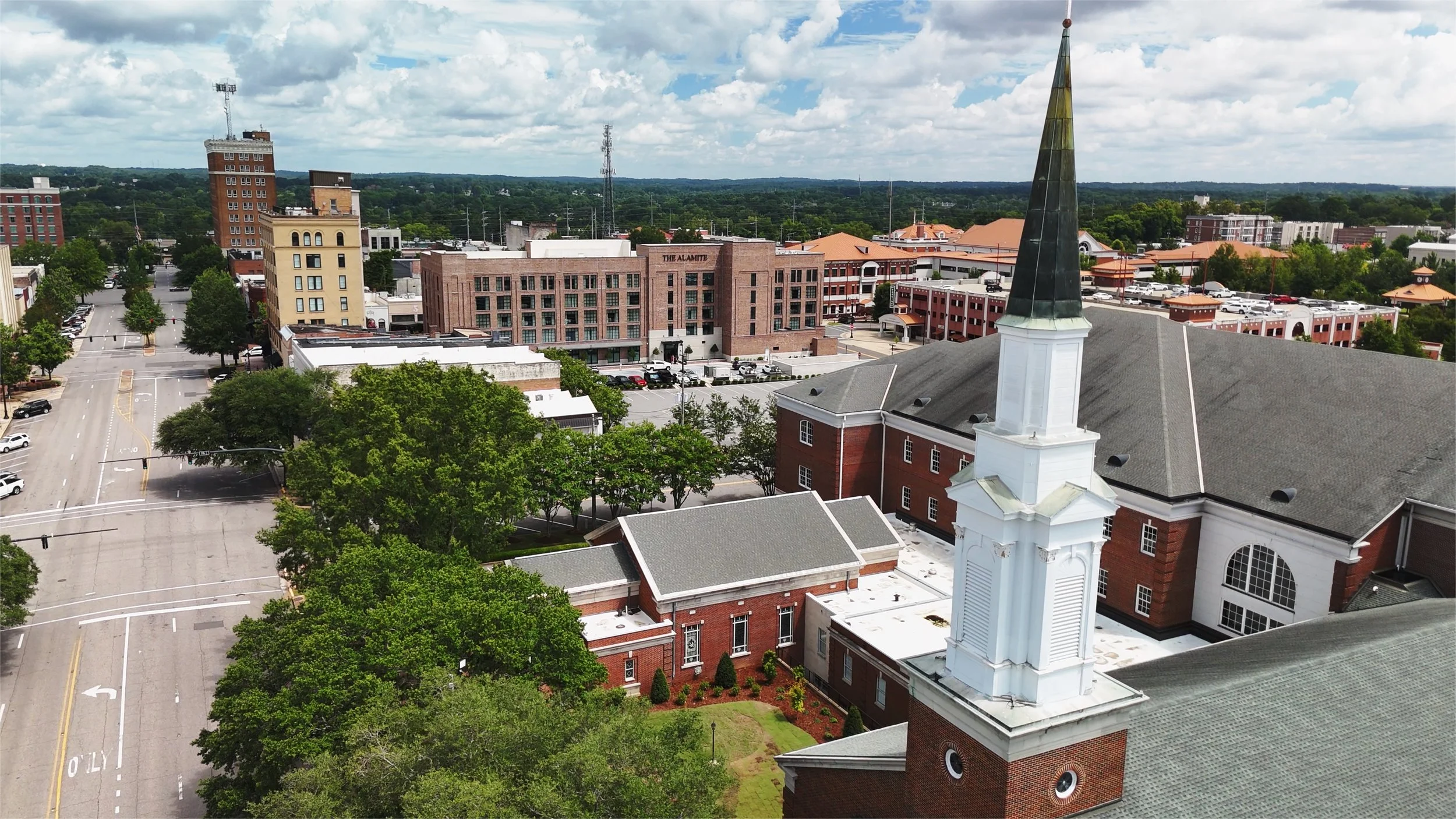 A cityscape with a church steeple in the foreground, surrounded by trees, and downtown buildings in the background under a partly cloudy sky.