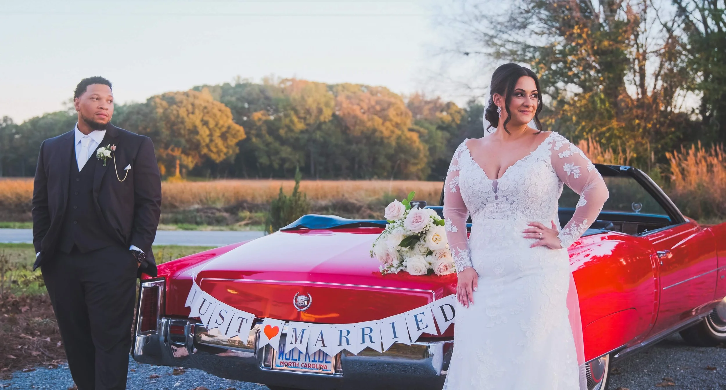 A newlywed couple stands next to a red vintage convertible car with a 'Just Married' banner in North Carolina. The bride wears a lace wedding dress with long sleeves, and the groom is dressed in a black tuxedo with a boutonniere. The bride is holding a bouquet of white roses, and the groom is leaning against the car, looking at her. The background features trees and a clear sky during sunset.