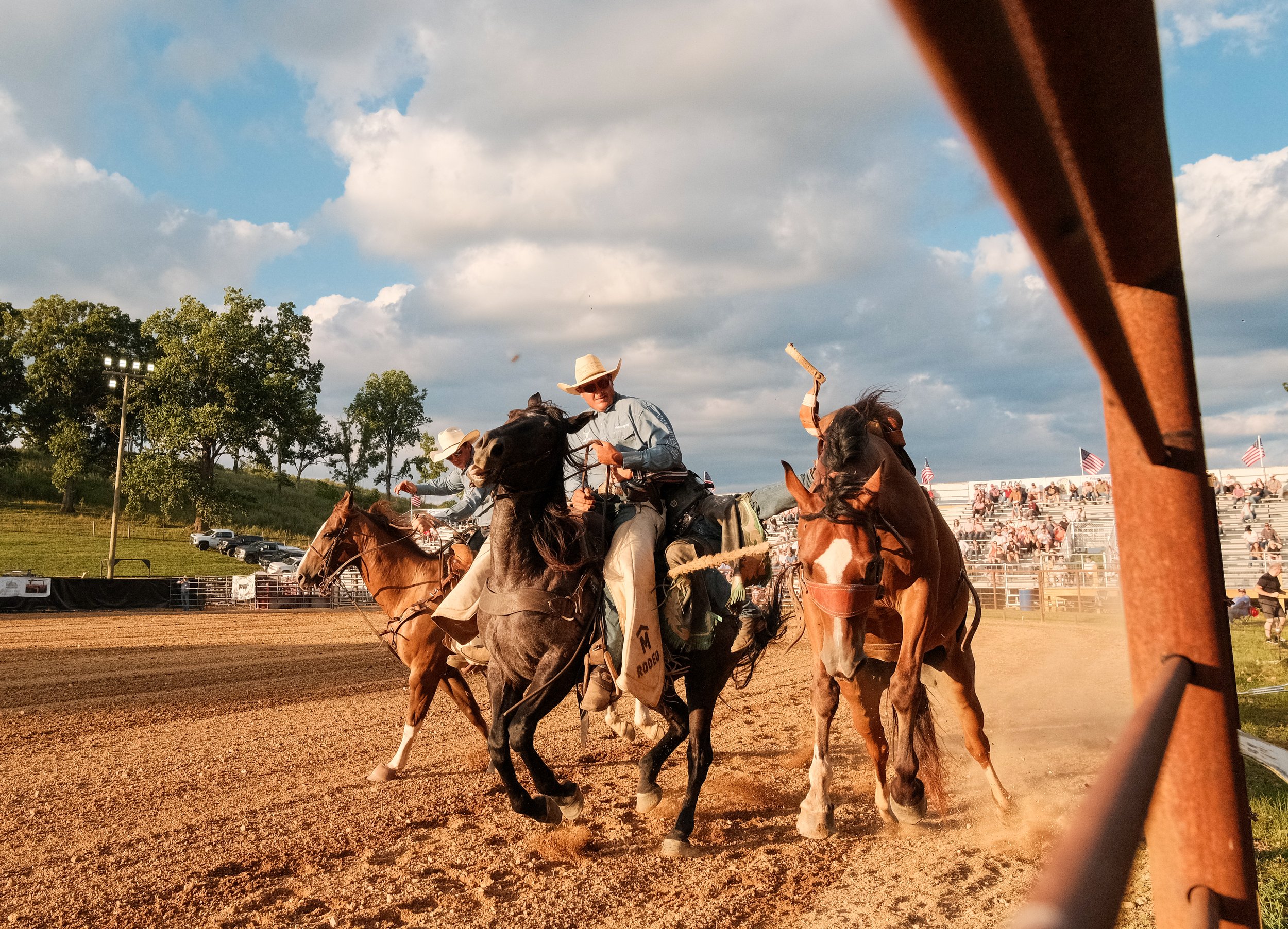 Cowboys participating in a rodeo event, riding horses on a dirt arena with spectators and American flags in the background.