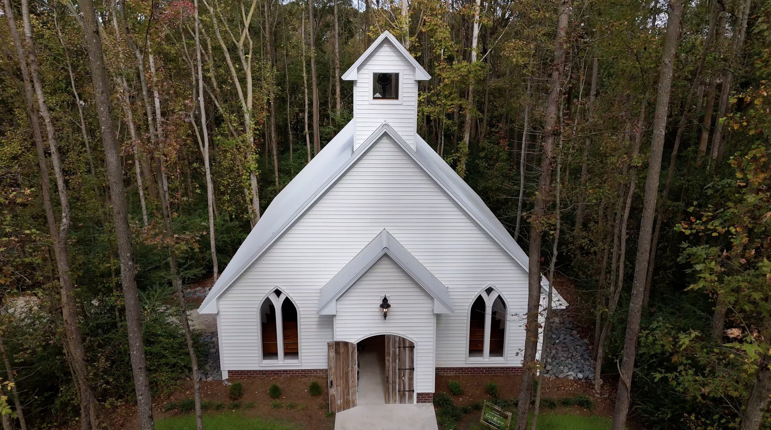 A small white church with a steep roof and arched windows, surrounded by trees. The church has a bell tower and wooden doors.