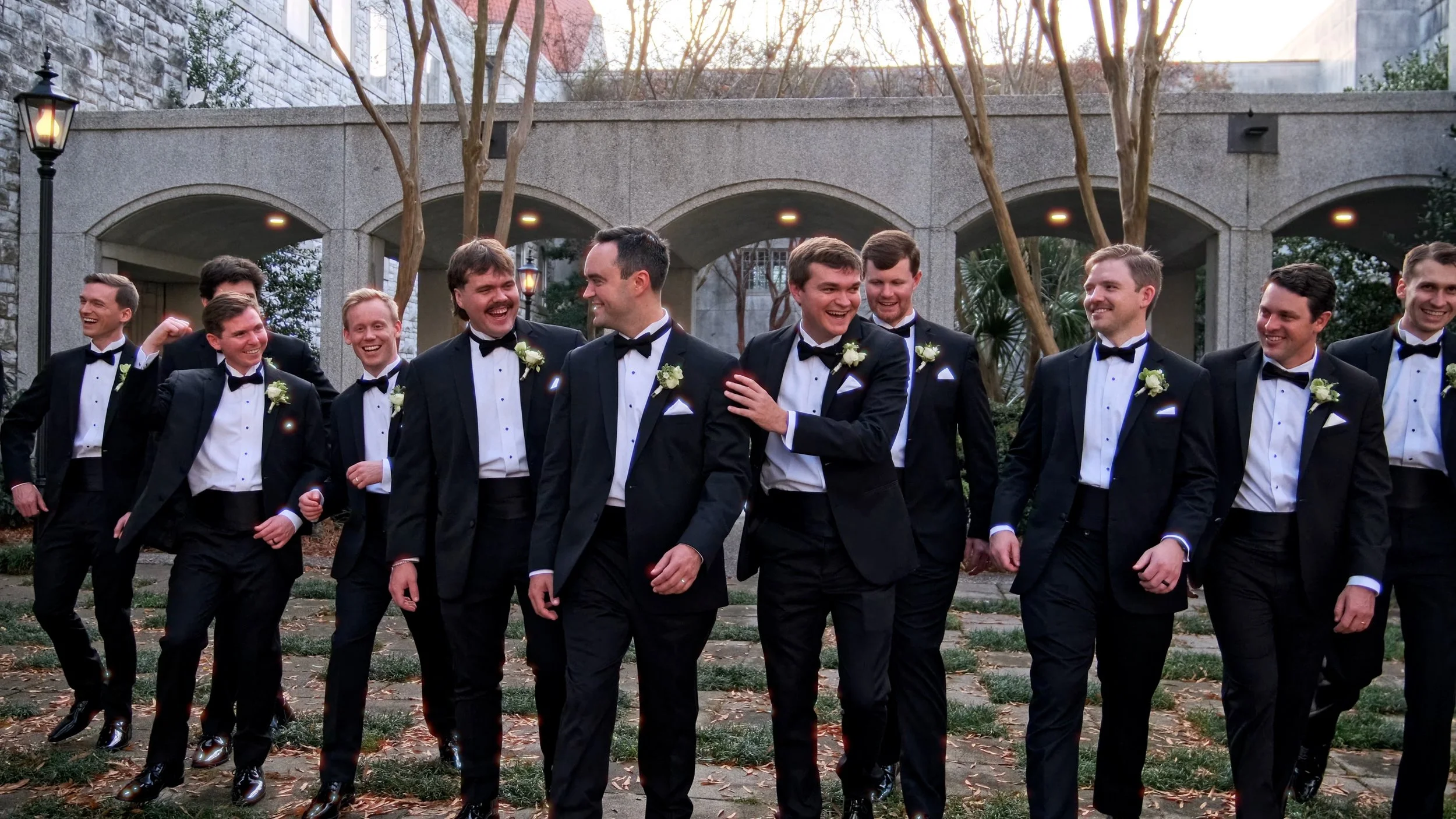 Group of groomsmen in tuxedos walking outdoors and smiling at each other.