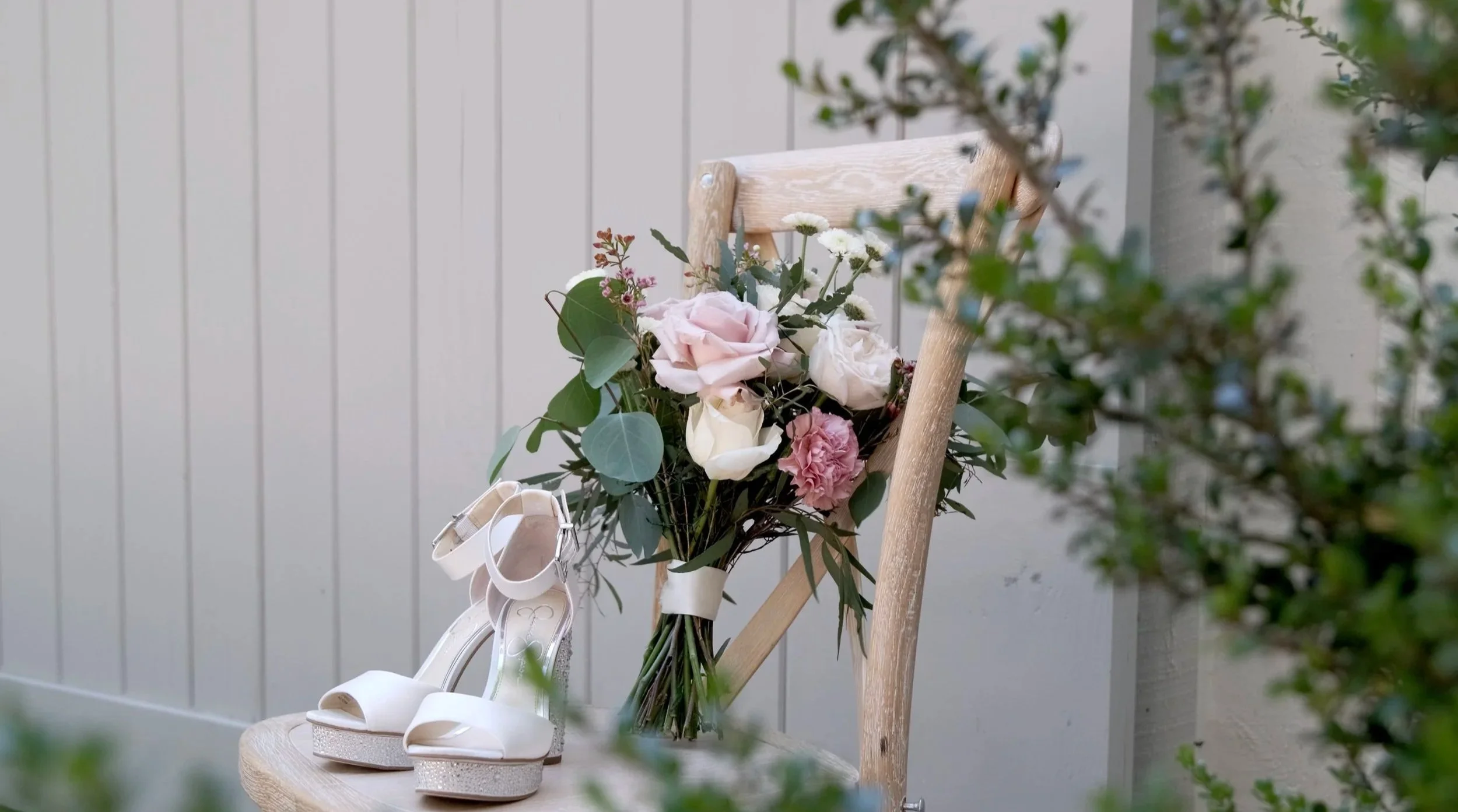White high-heeled sandals with ankle straps resting on a wooden surface next to a bouquet of pink and white roses with green leaves, placed on a wooden chair.