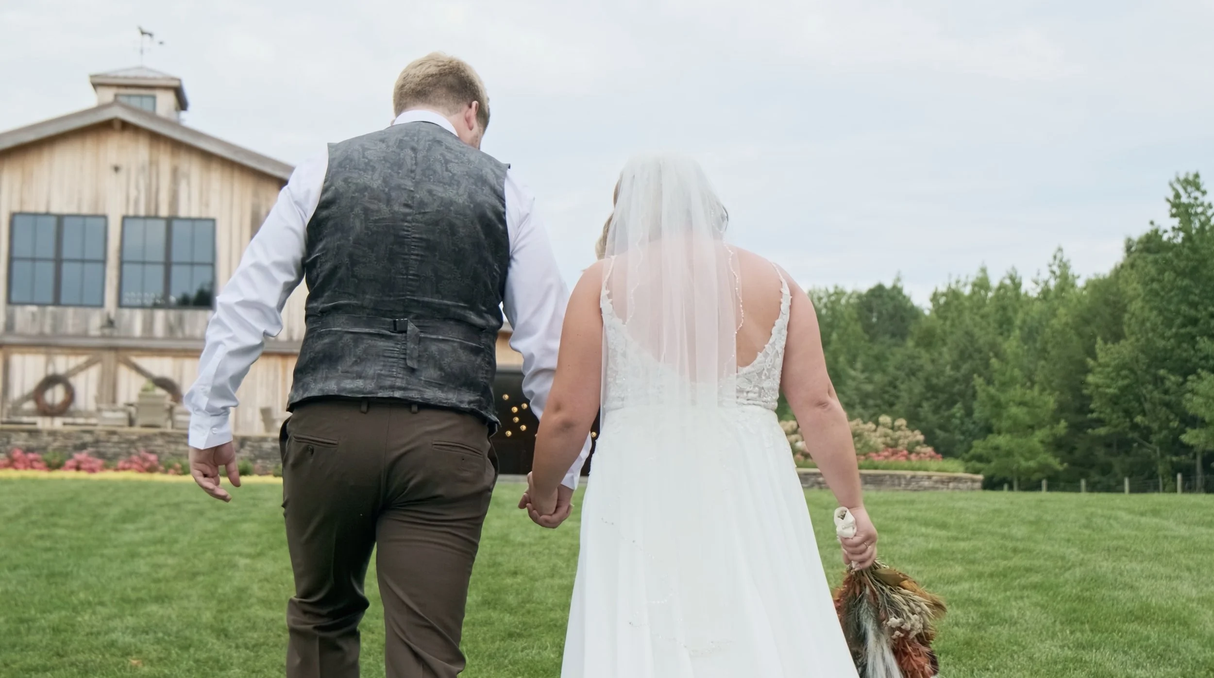 A bride and groom walking hand in hand on a grassy lawn, with a rustic barn and green trees in the background, during a wedding.