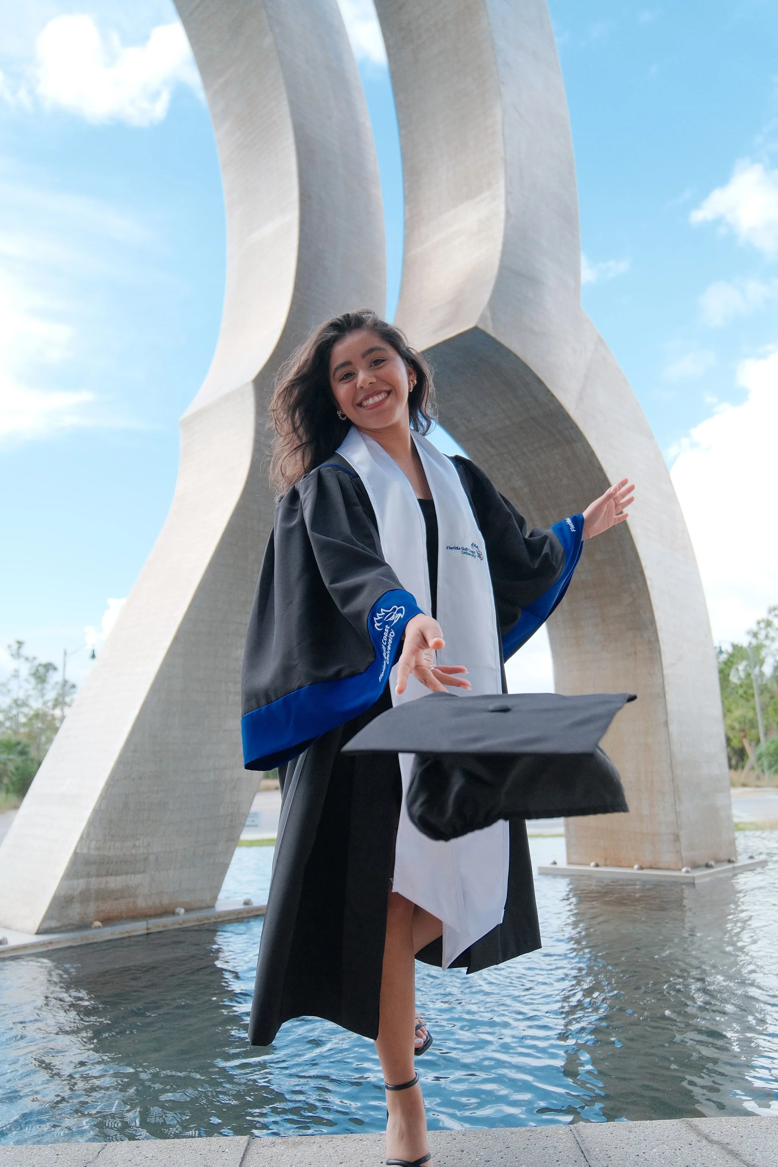 A young woman in a graduation gown and cap standing outdoors, tossing her cap in front of a large metal sculpture and water feature, with a partly cloudy sky above.