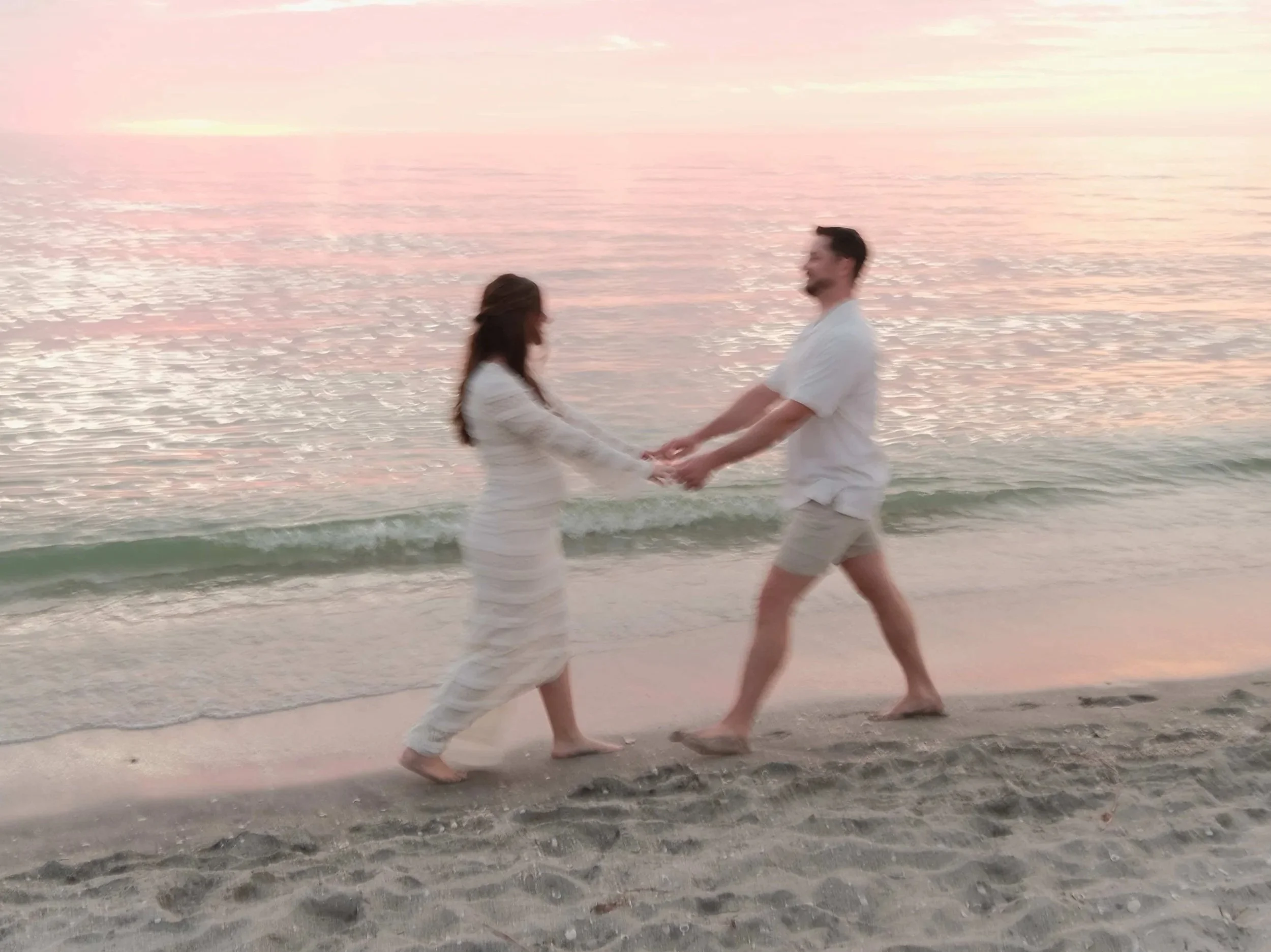 A couple holding hands and facing each other on the beach at sunset, with pink and orange sky reflected in the water.
