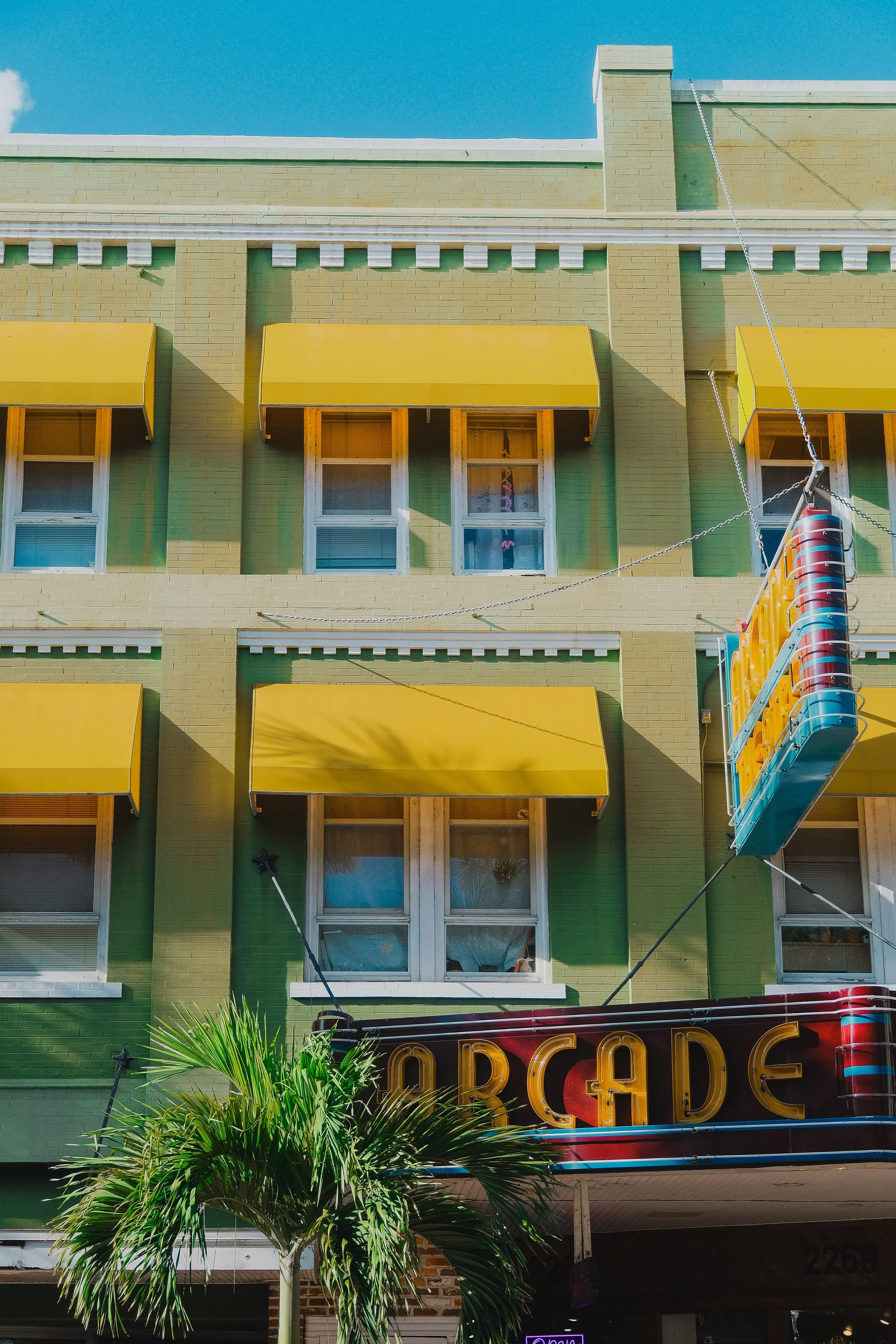 A colorful building with green bricks, yellow awnings over windows, and a neon sign that says 'ARCADE.' There is a palm tree in the foreground and a clear blue sky above.
