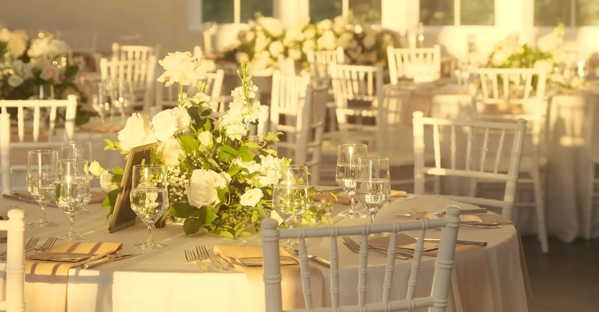 Elegant banquet table set for a wedding or special event with white floral centerpiece, glassware, silverware, and white chairs, illuminated by warm sunlight.