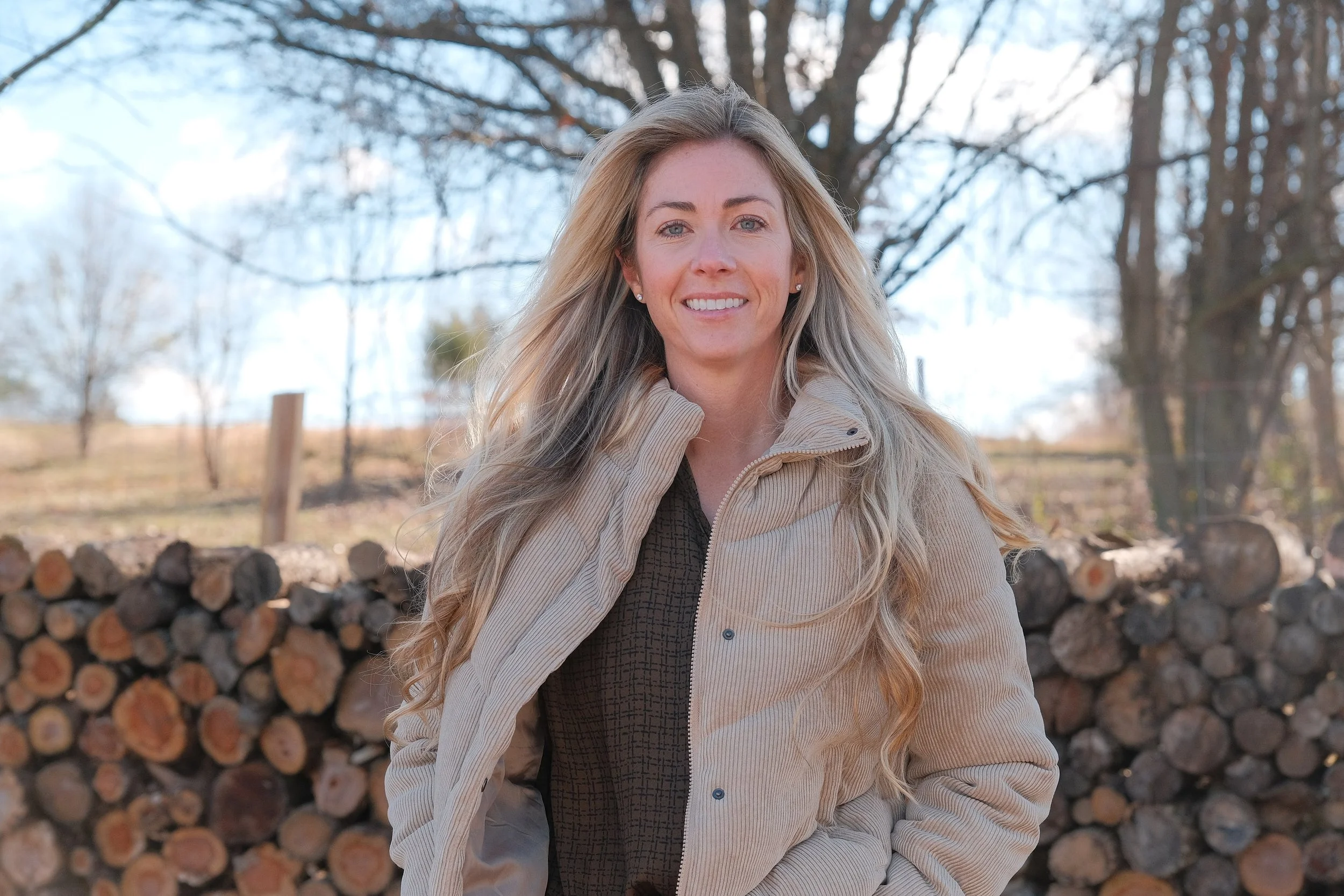 A woman with long blonde hair standing outdoors in front of a pile of logs, wearing a beige jacket and smiling.