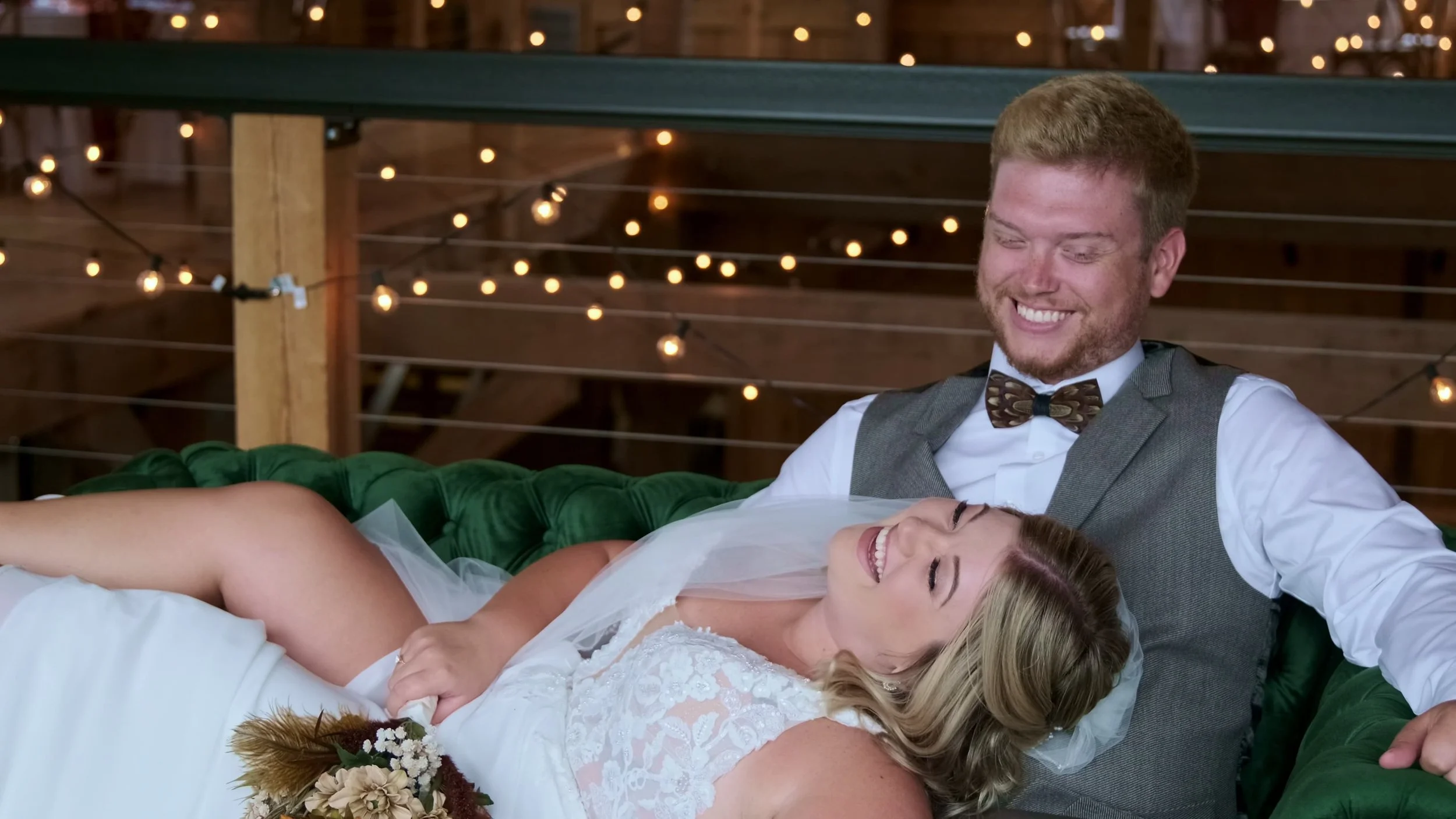A happy bride and groom sitting on a green velvet sofa, with string lights hanging in the background, during their wedding celebration.