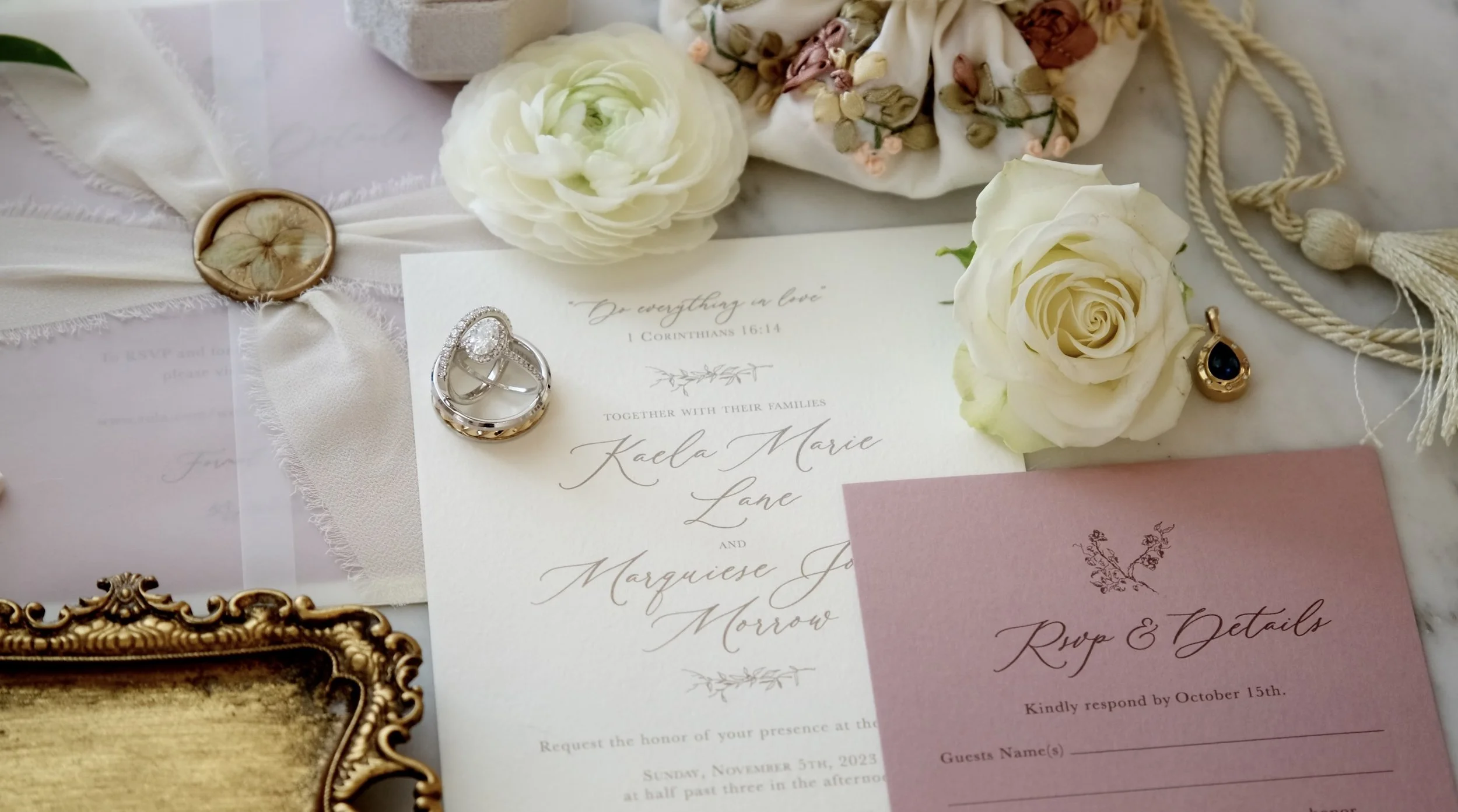 Wedding invitation surrounded by white roses, a ring, a gold pendant, patterned fabric, and a pink RSVP card on a white surface.