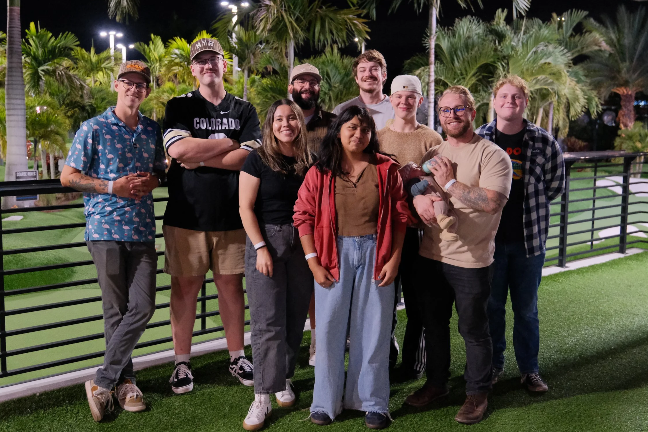 A group of ten people standing outdoors at night near a black railing, with palm trees and green artificial grass in the background, smiling for the camera.