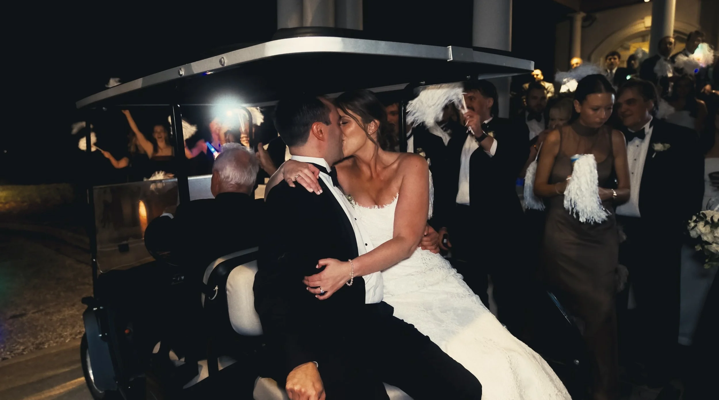 A bride and groom kissing on a golf cart during their wedding celebration, surrounded by guests in formal attire at night.