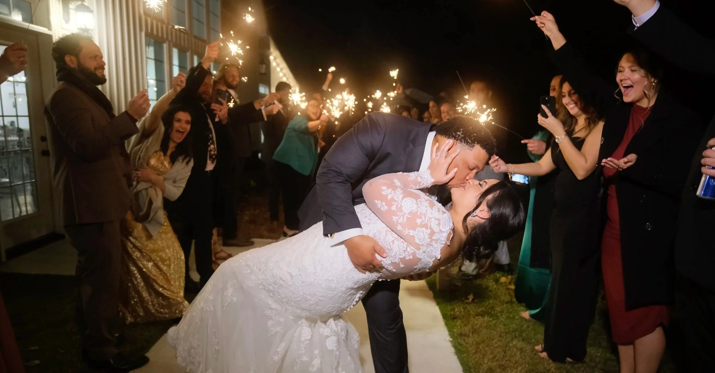 A newlywed couple shares a kiss during their wedding reception, surrounded by guests holding sparklers in the evening outside a building.
