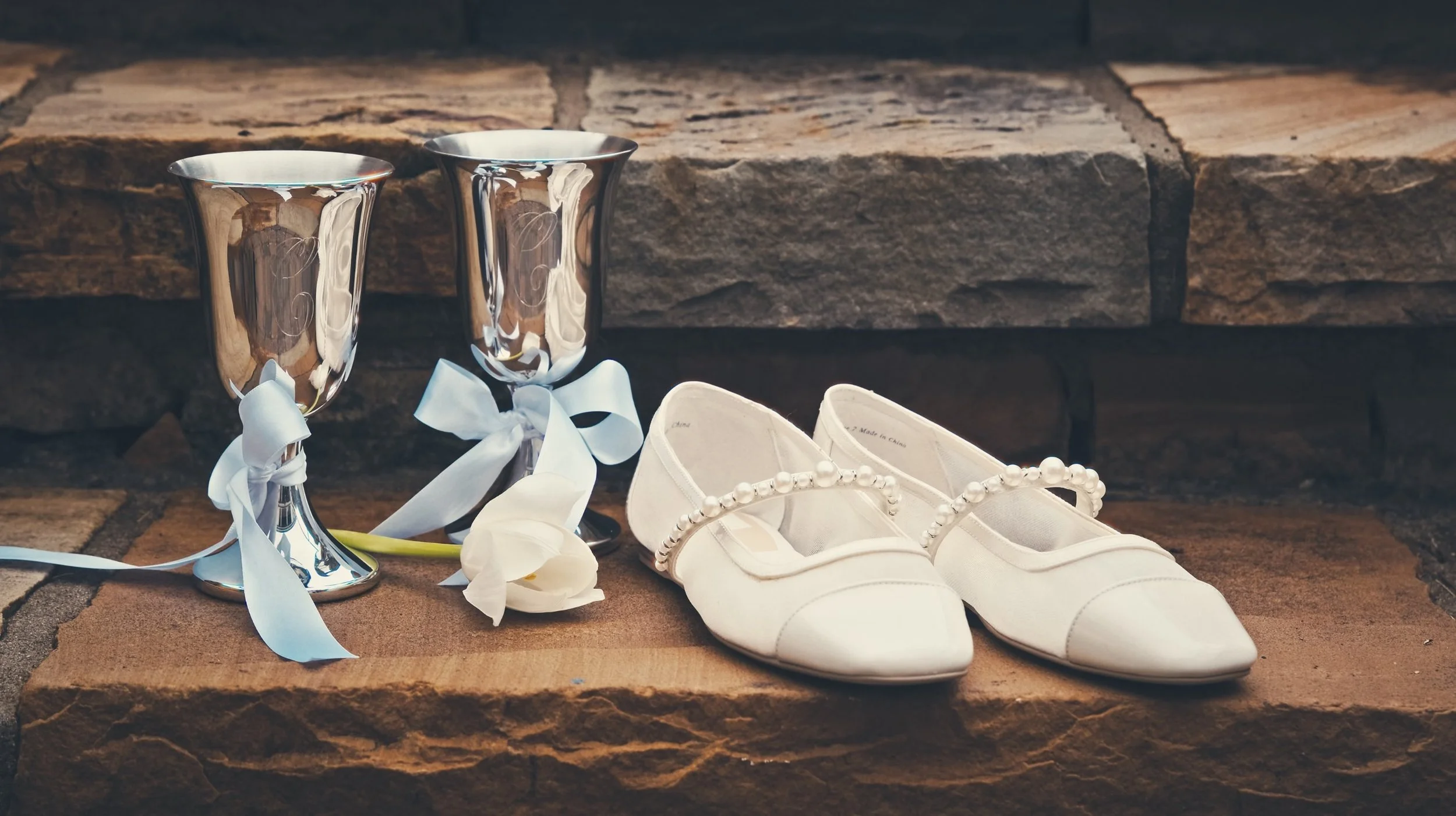 Silver chalices with white ribbons, white ballet flats with pearl accents, and white calla lilies placed on a stone ledge.